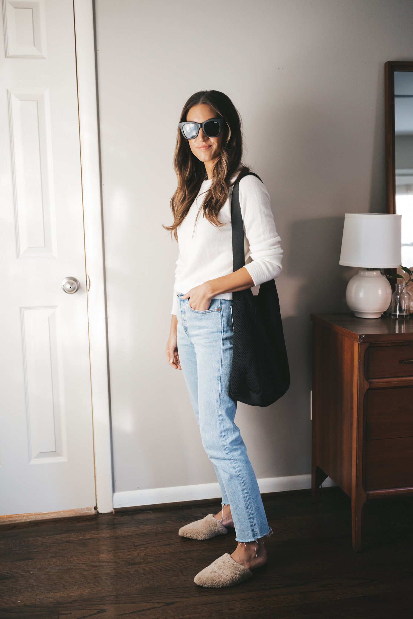 ashley wearing a cream colored crewneck sweater, light wash jeans, and tan shearling mules with a black tote bag and black square frame sunglasses