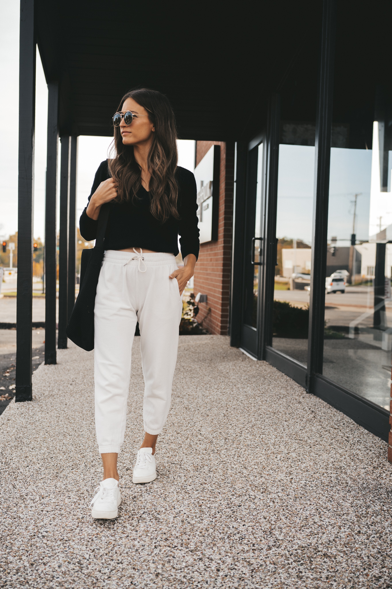 ashley wearing a black v-neck cashmere sweater, white joggers, white sneakers, a black mesh tote bag, and gold frame round sunglasses standing next to a brick building with black windows