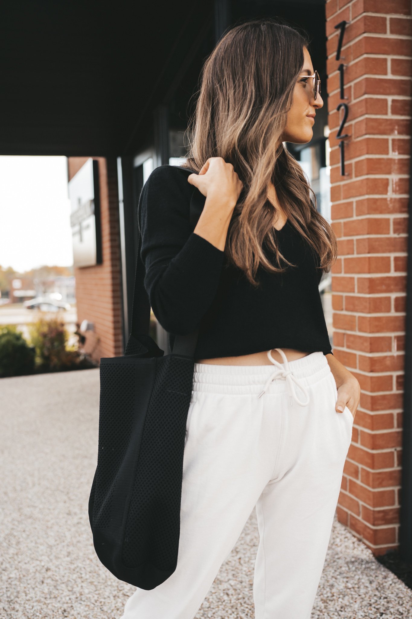 ashley wearing a black v-neck cashmere sweater, white joggers, white sneakers, a black mesh tote bag, and gold frame round sunglasses standing next to a brick building with black windows