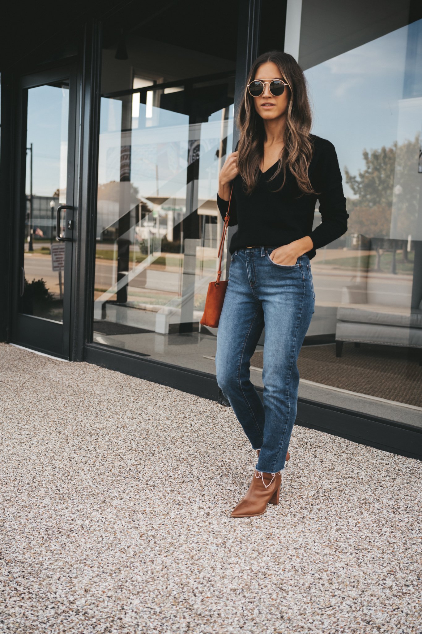 ashley wearing a black v-neck cashmere sweater, medium wash mom jeans, cognac heeled booties, and a dark red crossbody bag bag, and gold frame round sunglasses standing next to a building with black windows