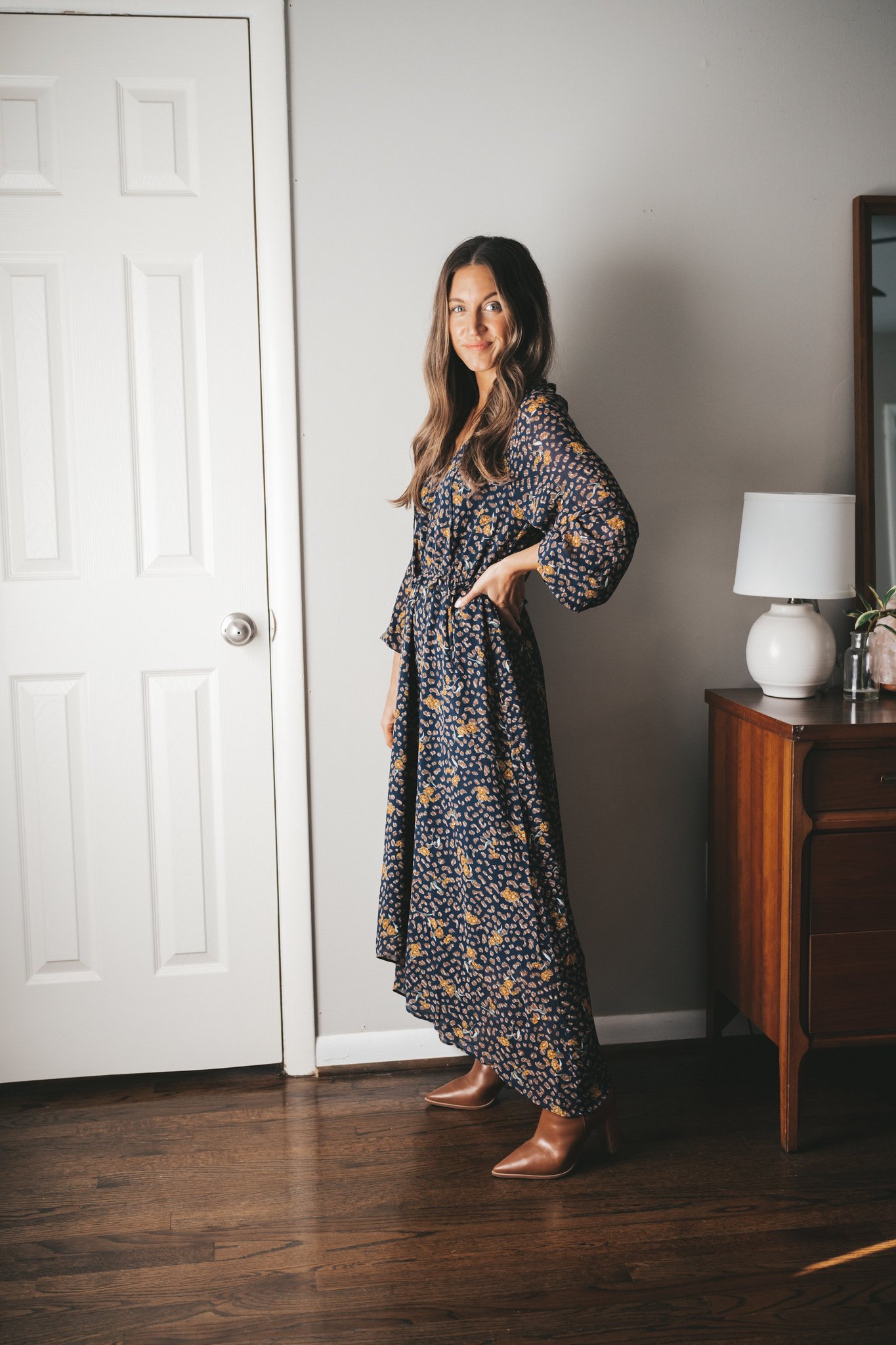 ashley wearing a navy floral maxi dress with heeled cognac booties standing next to a vintage wood dresser with a white table lamp