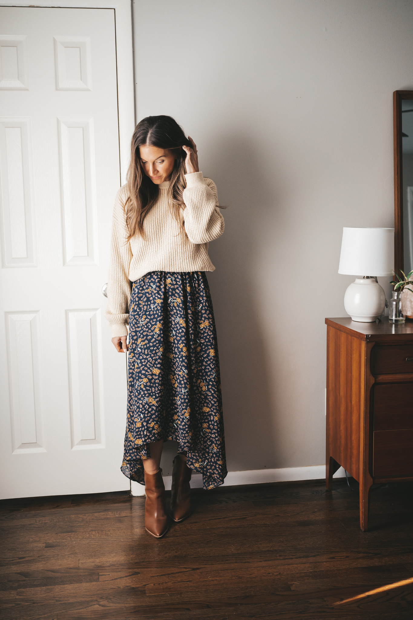 ashley wearing a navy floral maxi dress with a cream colored sweater over it with heeled cognac booties standing next to a vintage wood dresser with a white table lamp
