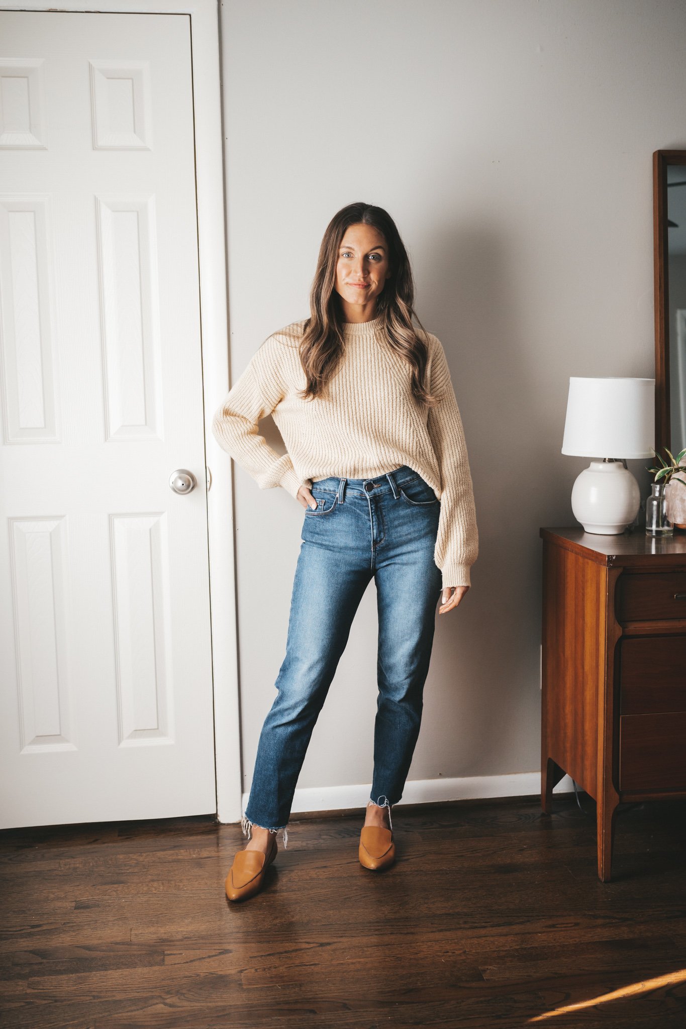 ashley wearing a cream colored sweater with mid-wash mom jeans, and cognac loafers standing next to a vintage wood dresser with a white table lamp