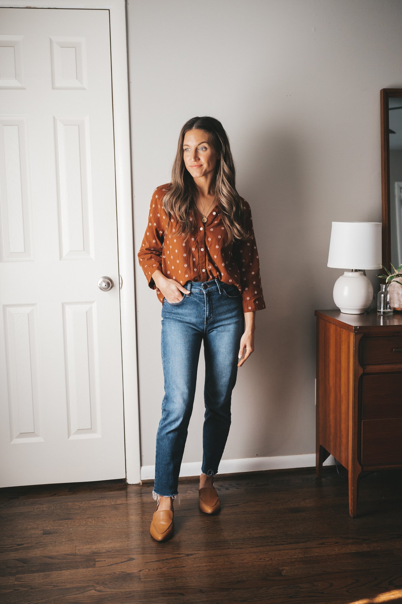 ashley wearing a patterned rust colored blouse, mid-wash mom jeans, and cognac loafers standing next to a vintage wood dresser with a whi