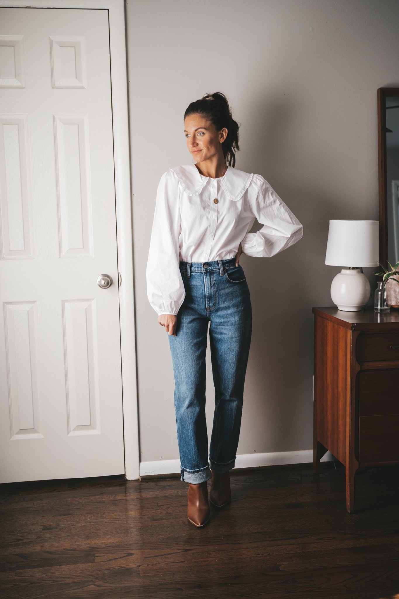 ashley wearing a white button down with a peter pan collar, straight leg denim cuffed, and cognac booties standing next to a vintage wood dresser with a white table lamp