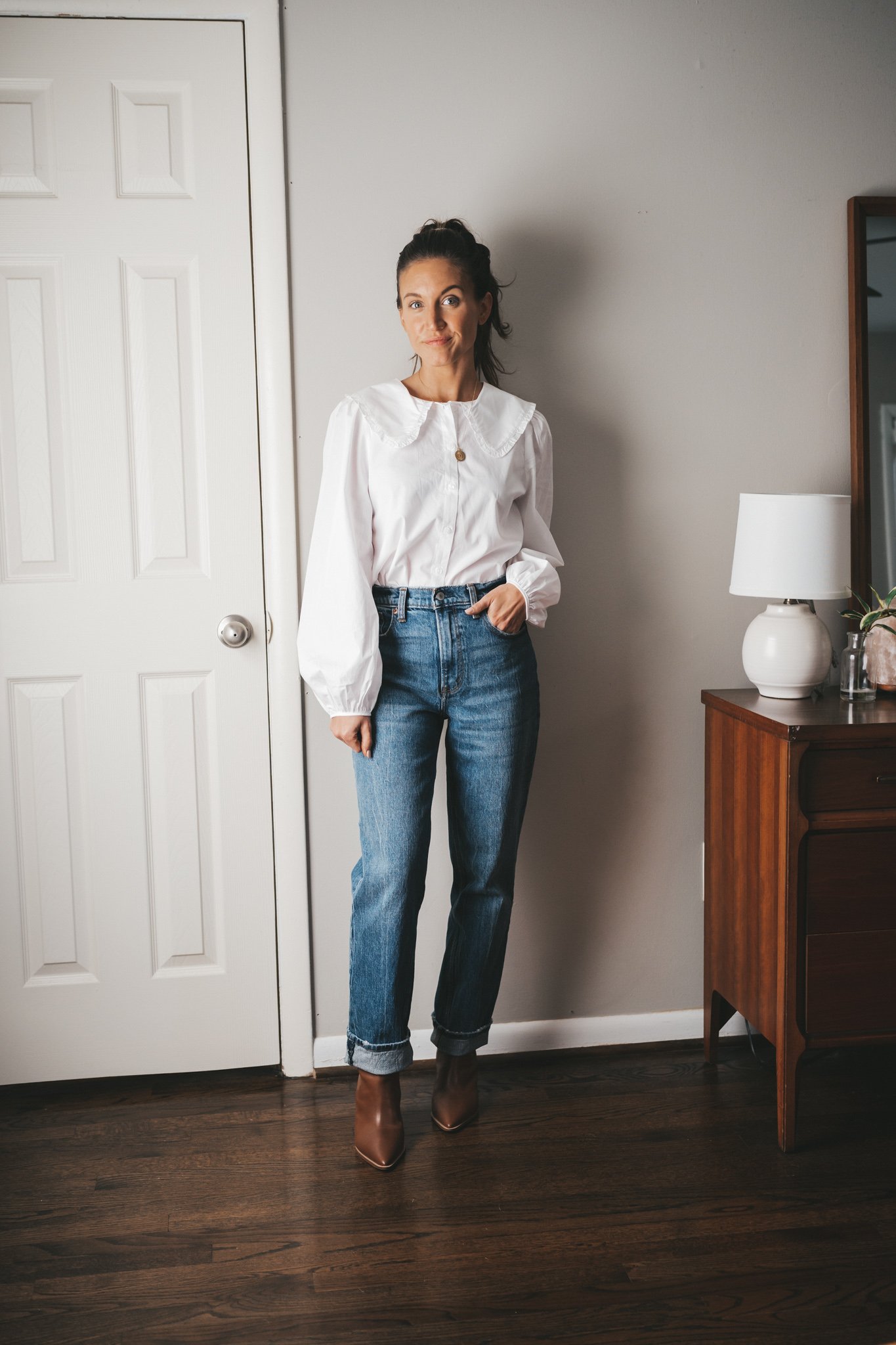 ashley wearing a white button down with a peter pan collar, straight leg denim cuffed, and cognac booties standing next to a vintage wood dresser with a white table lamp