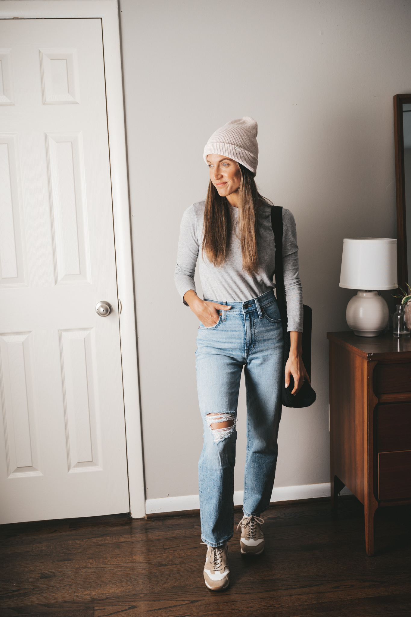 ashley wearing a gray tee, light wash straight distressed denim, tan and white sneakers, a white beanie and a black tote bag