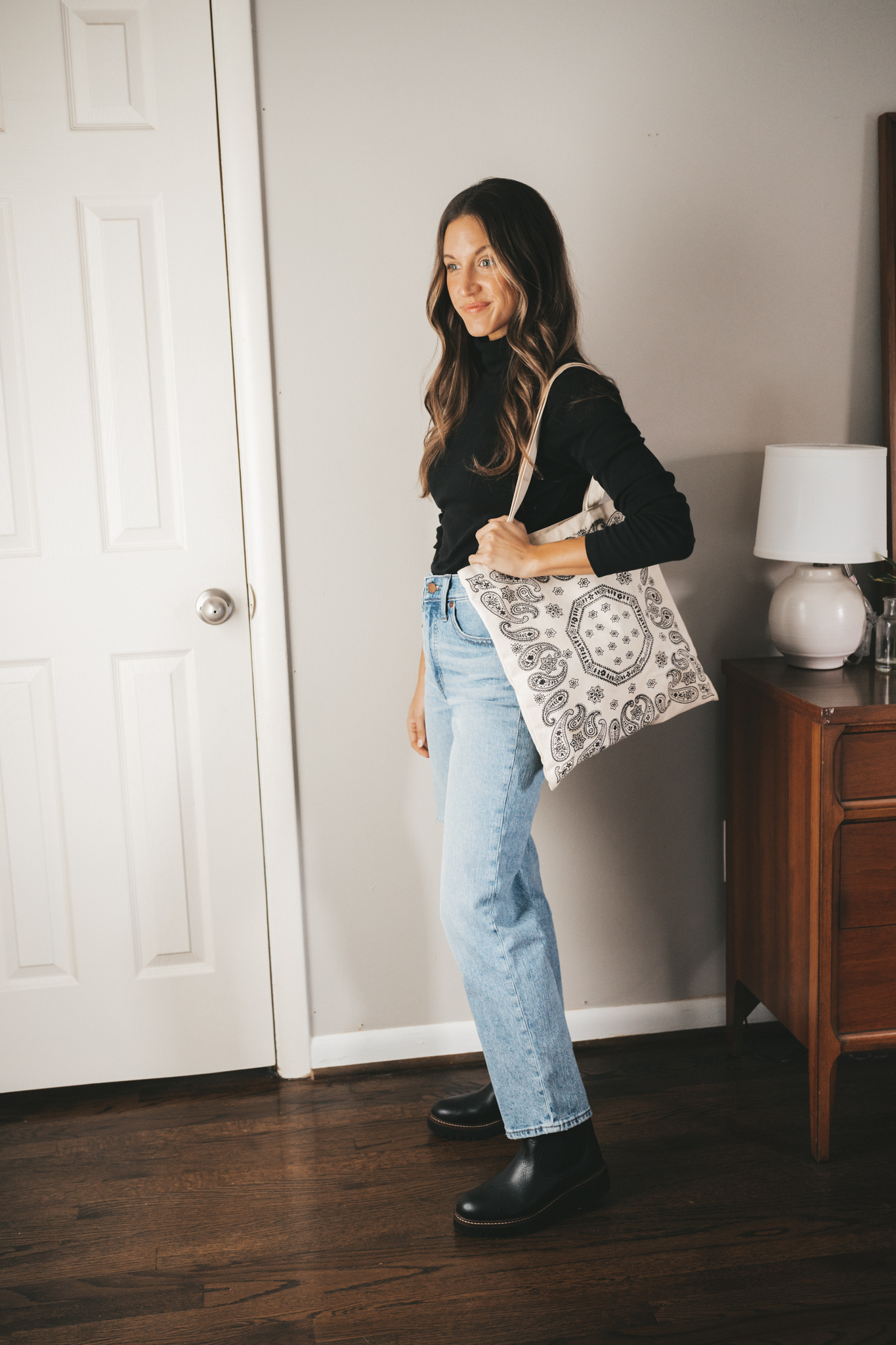 ashley wearing a black turtleneck, light wash distressed straight denim, black lug sole chelsea boots, and a canvas tote bag