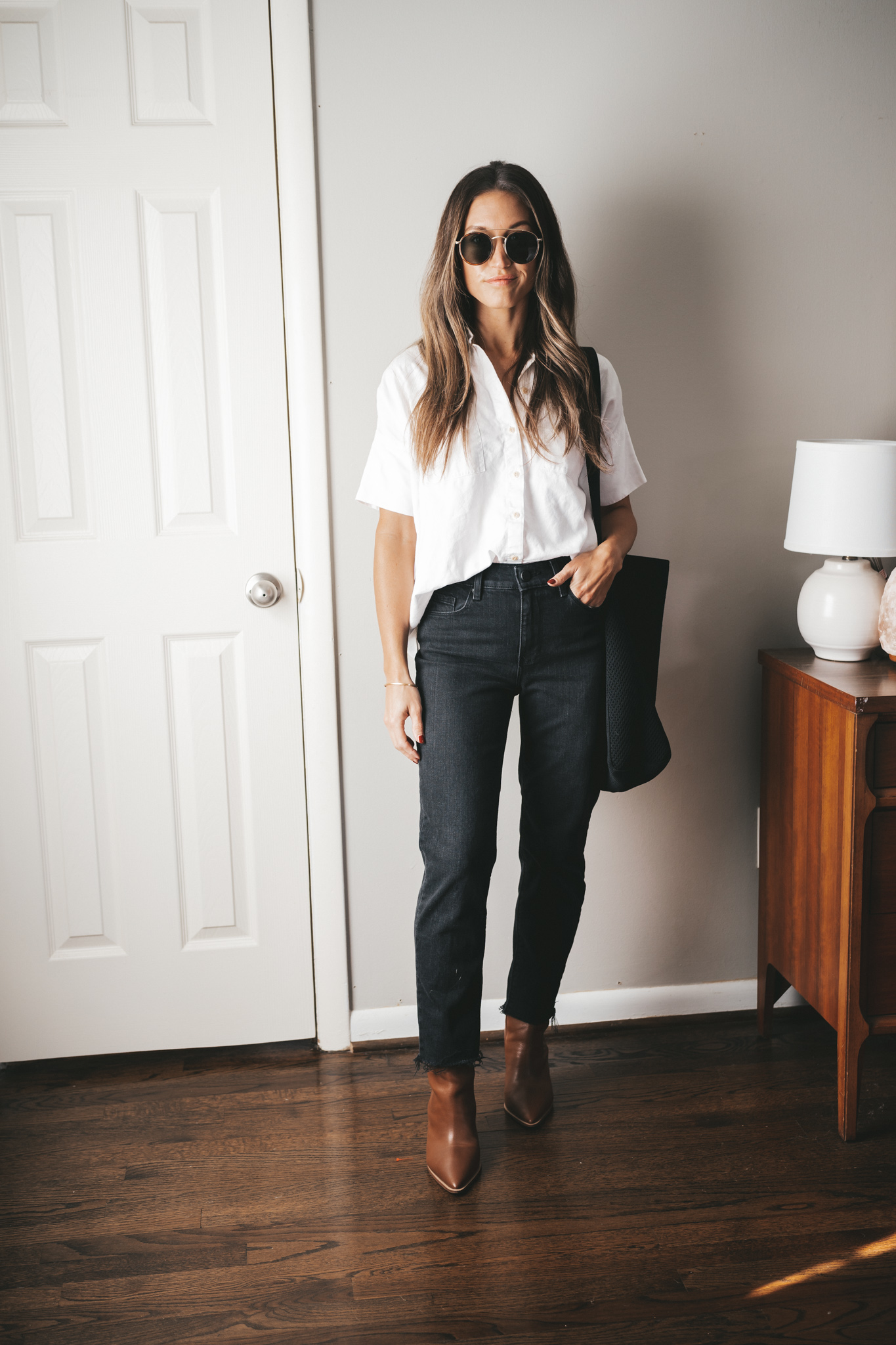 ashley wearing a boxy white button down, black mom jeans, brown heeled booties, a black tote bag, and gold frame round sunglasses