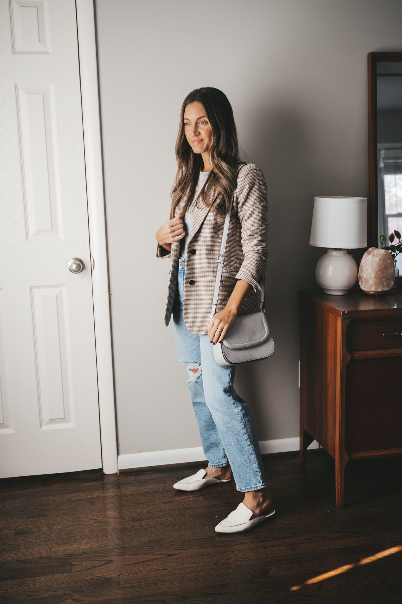ashley wearing a gray tee under a plaid blazer, light wash distressed staight denim, white mules, and a gray crossbody bag