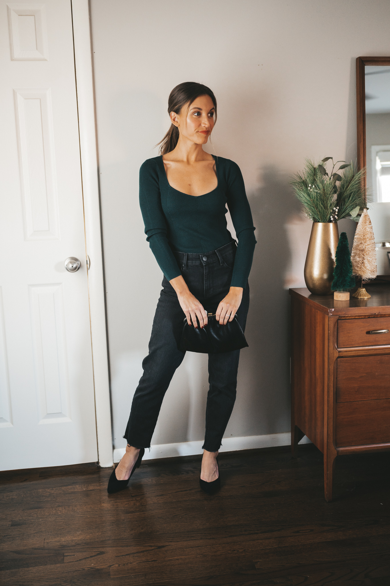 ashley wearing an emerald green square neck sweater with black denim and black block heeled pumps holding a vintage black clutch