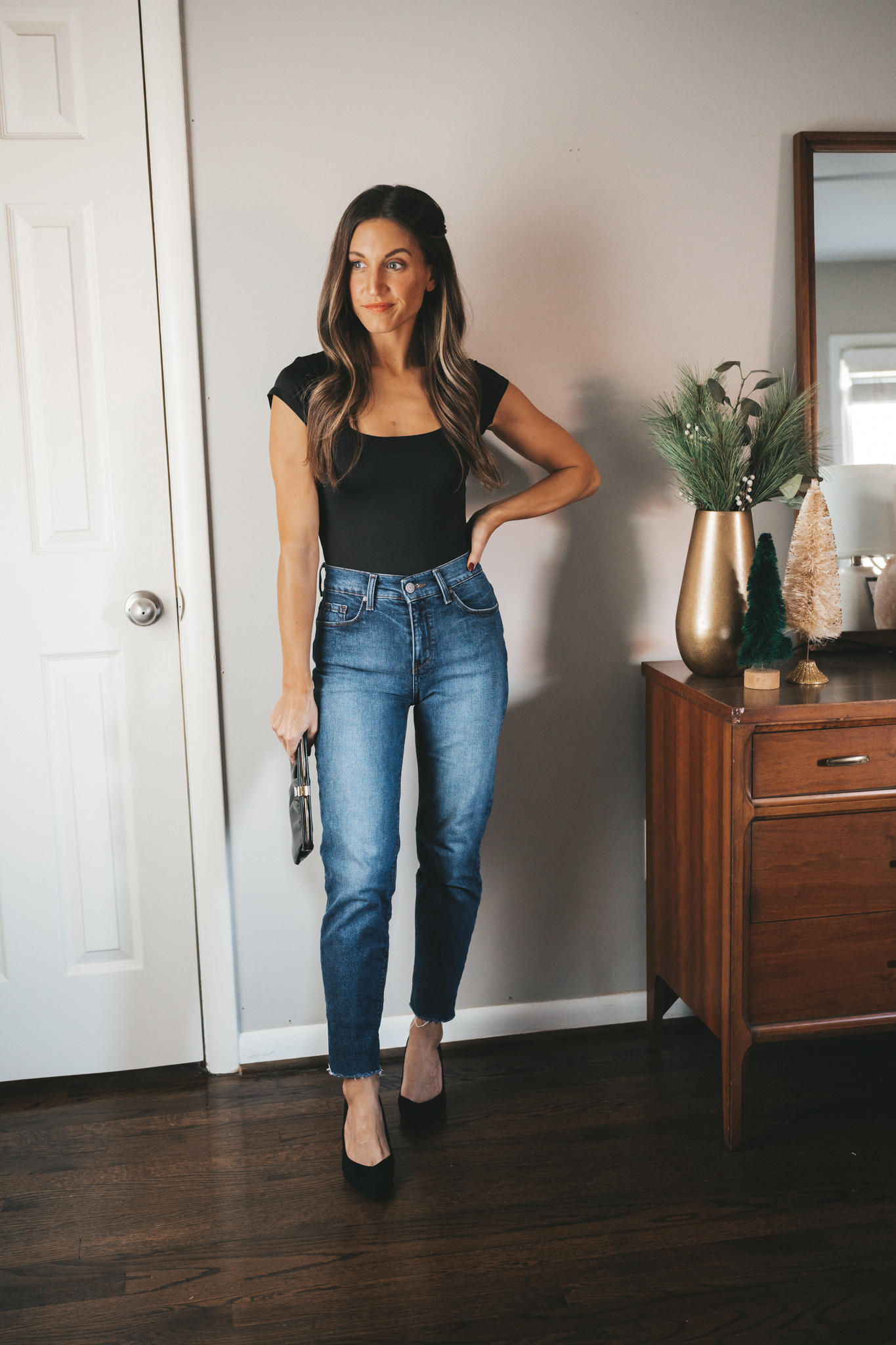 ashley wearing a black cap sleeve bodysuit with a square neck line, mid-wash jeans, and black block heeled pumps with a vintage black clutch