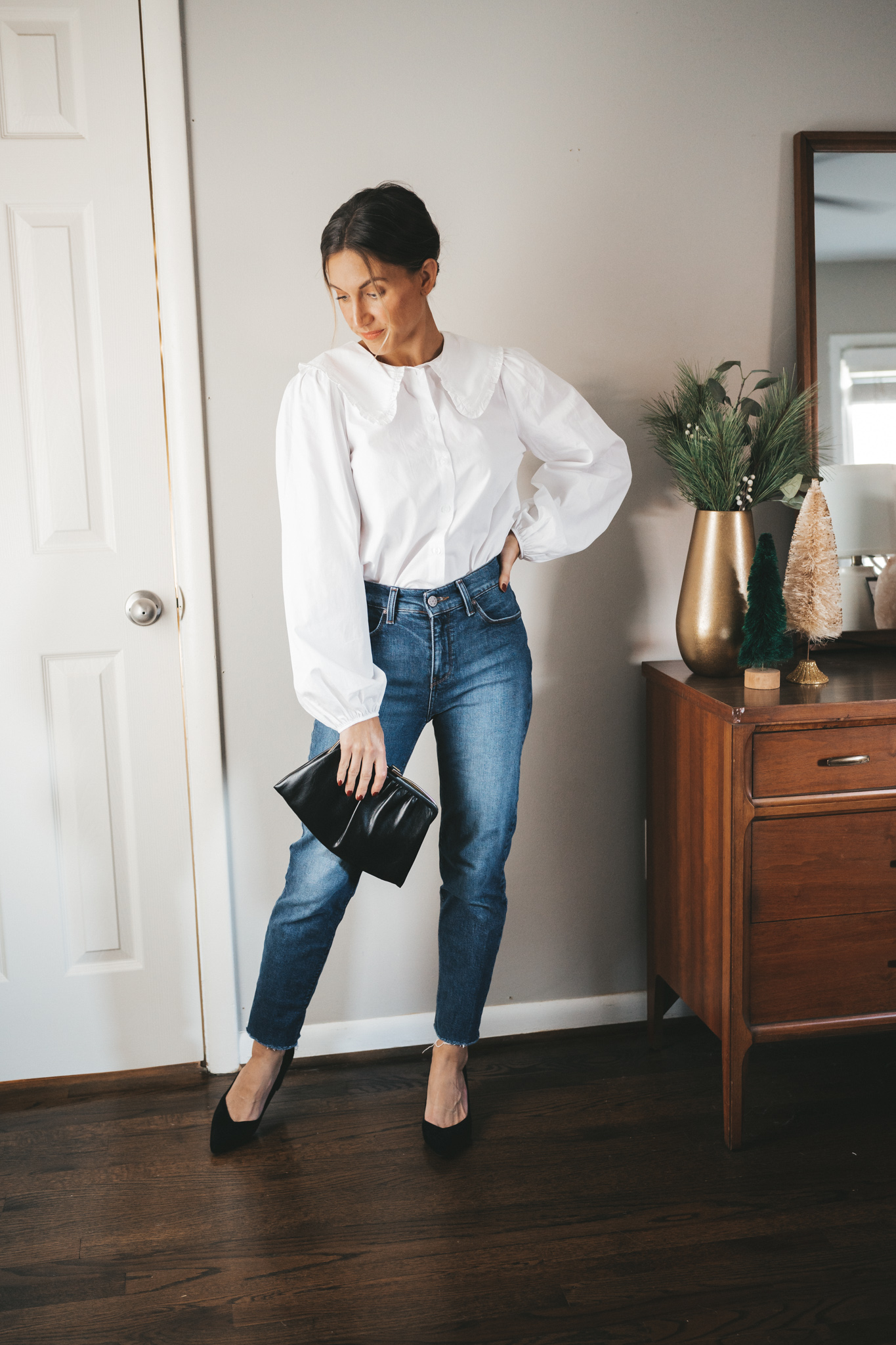 ashley wearing a white button down with a peter pan collar and balloon sleeves, mid-wash denim, black block heeled pumps, and a vintage black clutch with hair in a low bun