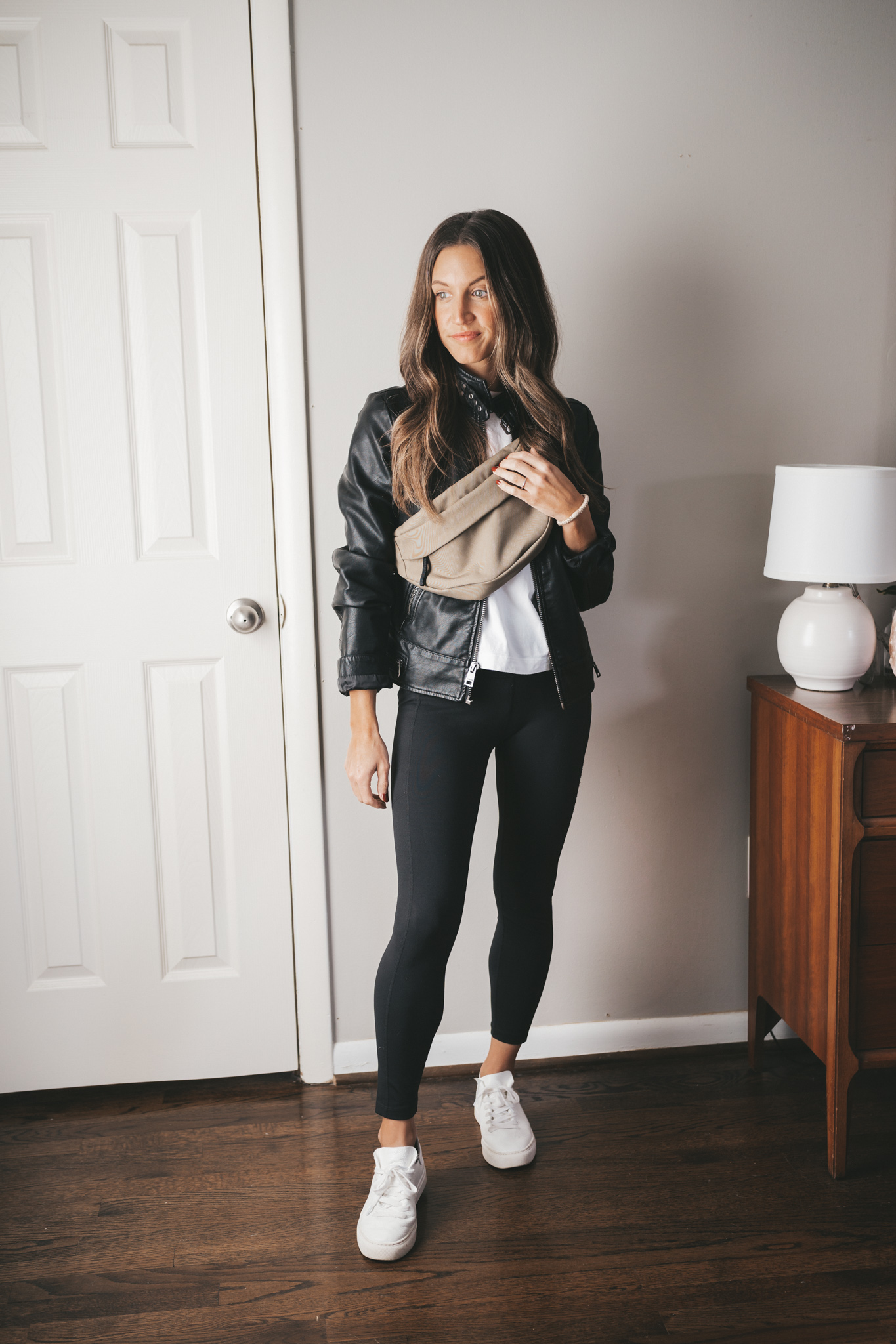 ashley wearing a white tee, faux leather moto jacket, black leggings, white sneakers, and an olive green belt bag worn over the shoulder
