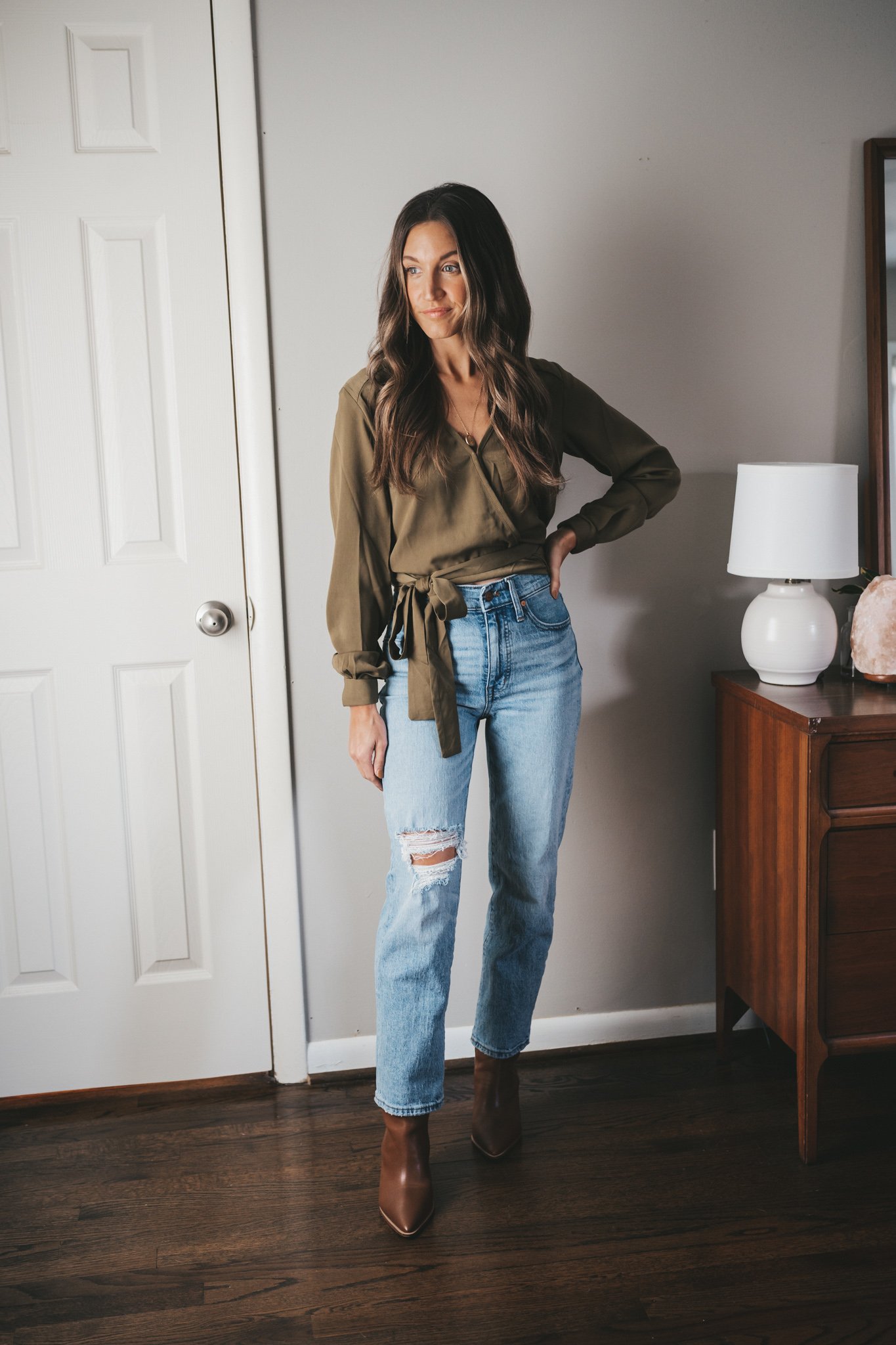 ashley wearing an olive green wrap top, light wash distressed denim, and brown ankle boots