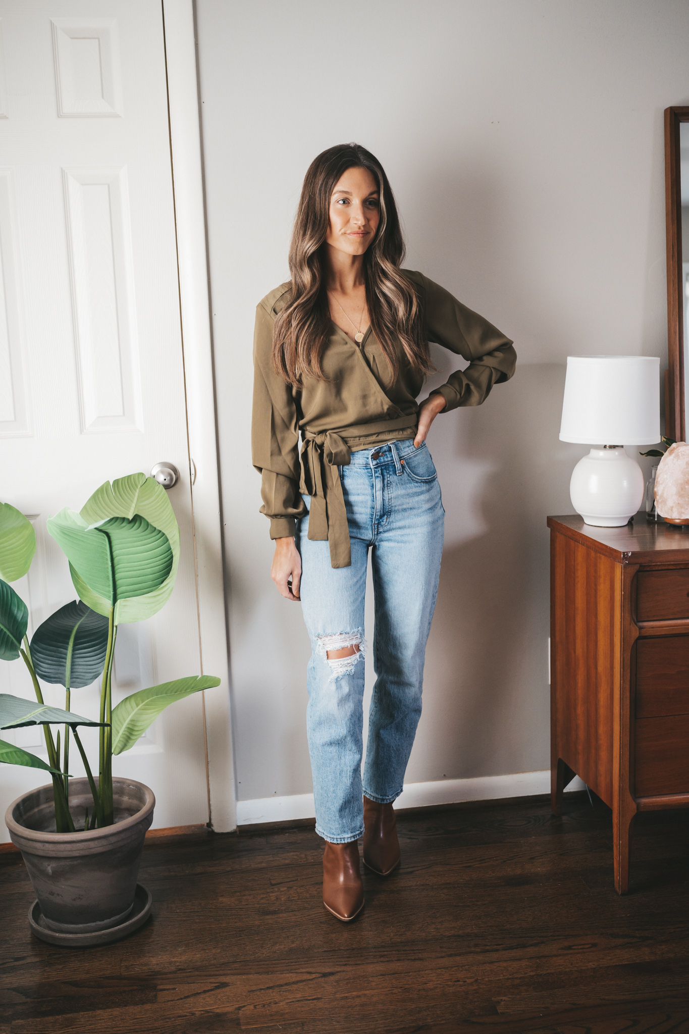 ashley wearing an olive green wrap top, light wash distressed denim, and brown ankle boots