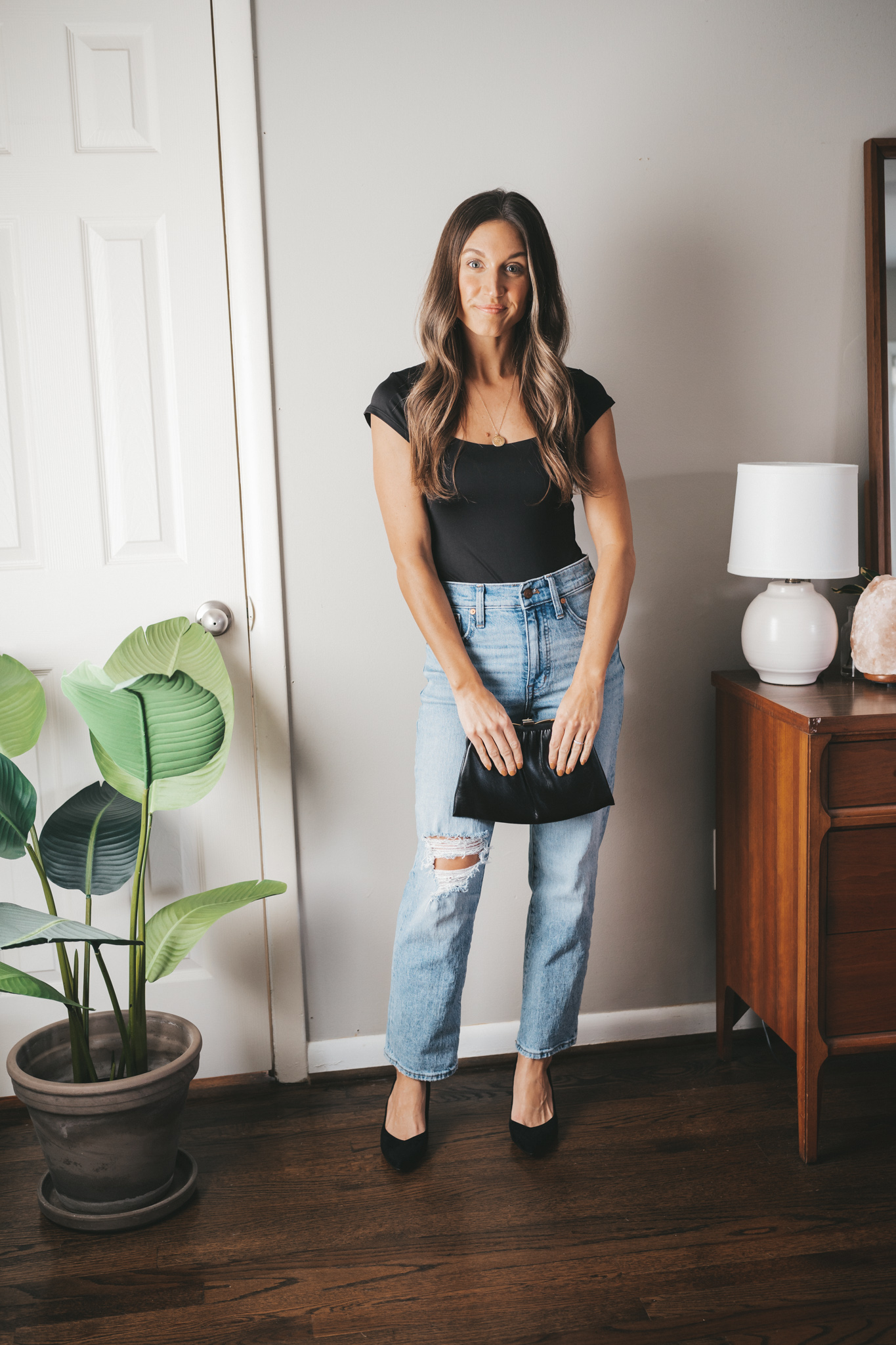 ashley wearing a black bodysuit, light wash distressed denim, black heels, and a black clutch