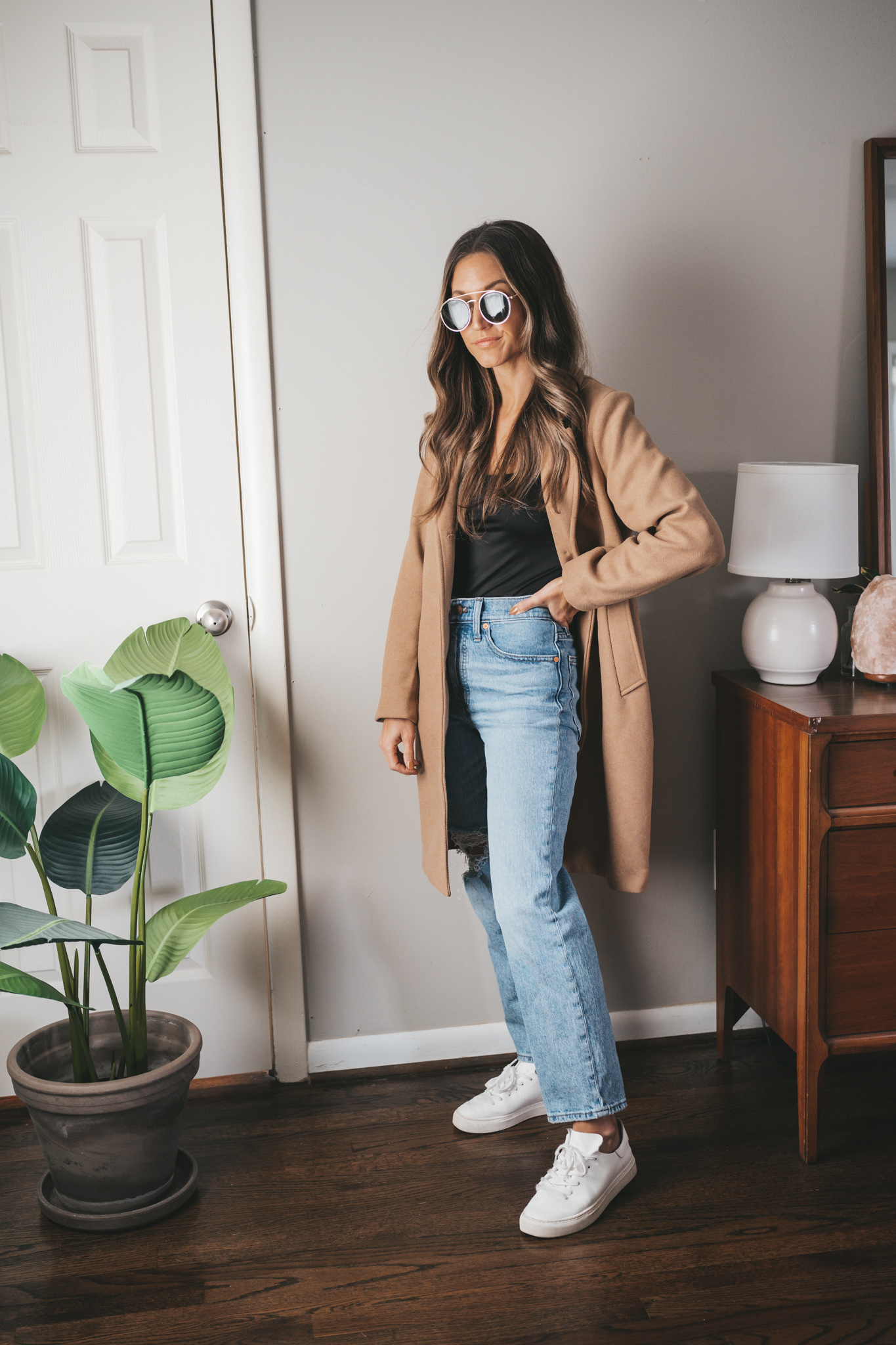 ashley wearing a black bodysuit under a camel coat, light wash distressed denim, white sneakers, and gold round frame sunglasses