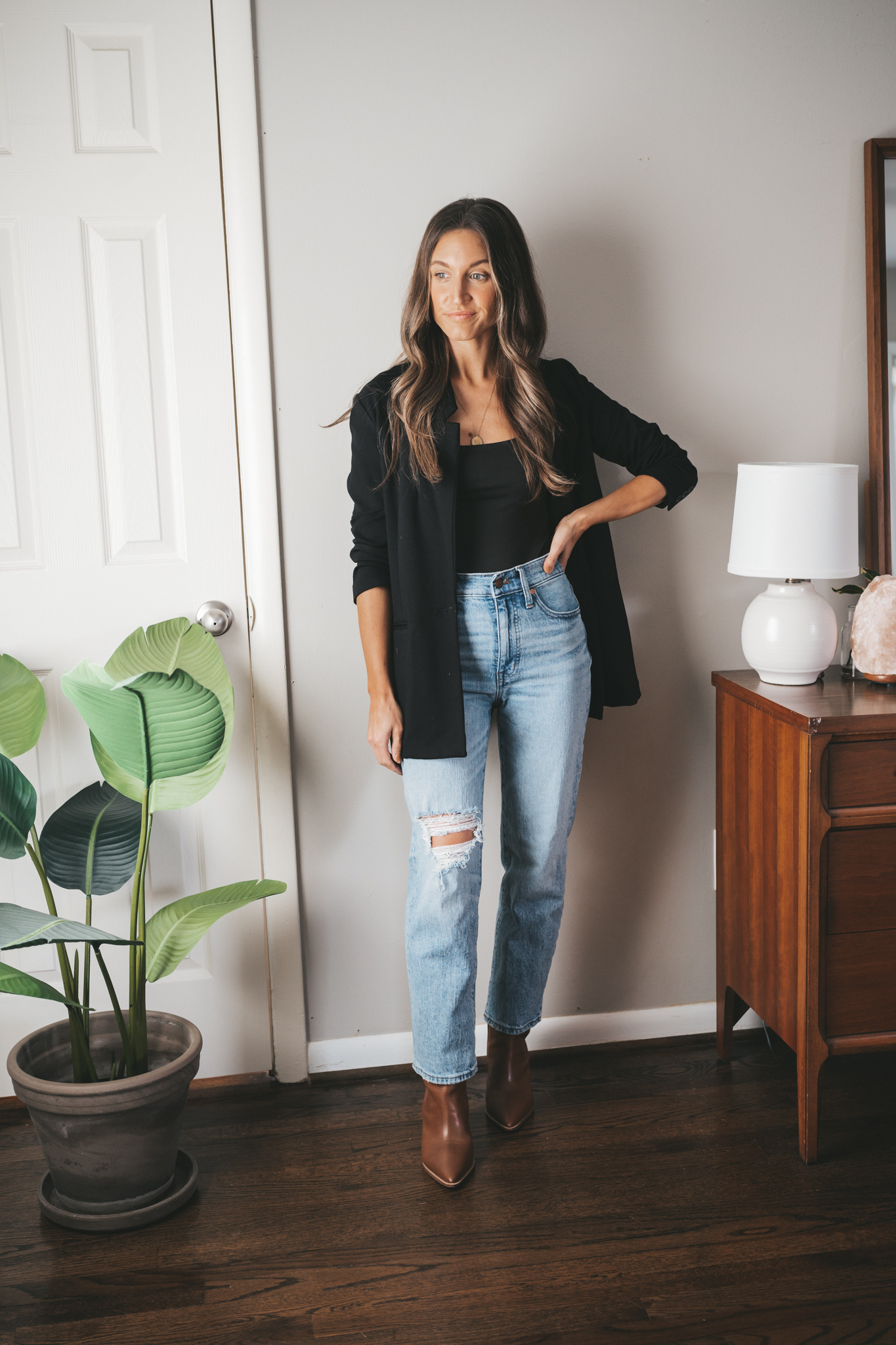 ashley wearing a black bodysuit under a black blazer, light wash distressed denim, and brown ankle boots