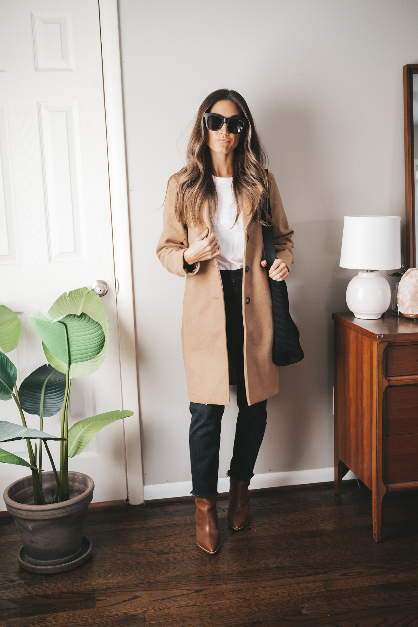 ashley wearing a white tee under a camel coat, black denim, brown ankle boots, and a black tote bag