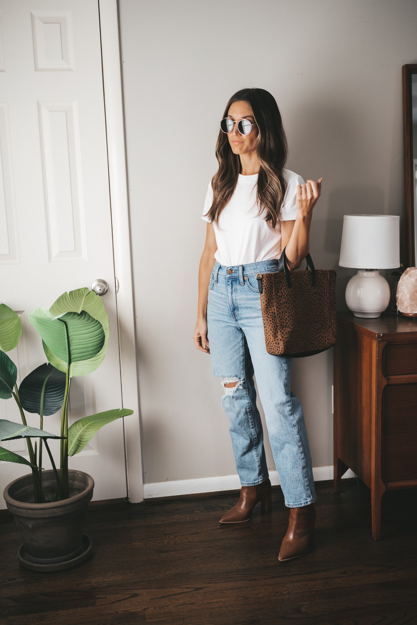 ashley wearing a white tee, light wash distressed denim, brown ankle boots, a leopard tote bag and gold round frame sunglasses