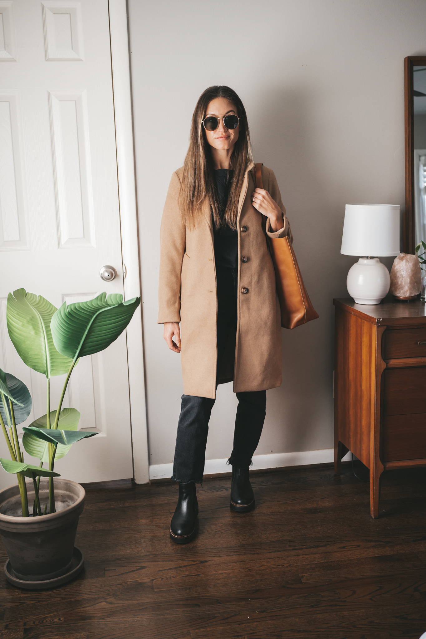 fall outfit idea with a black crewneck sweatshirt, black denim, camel coat, black chelsea boots, cognac tote bag and round gold frame sunglasses