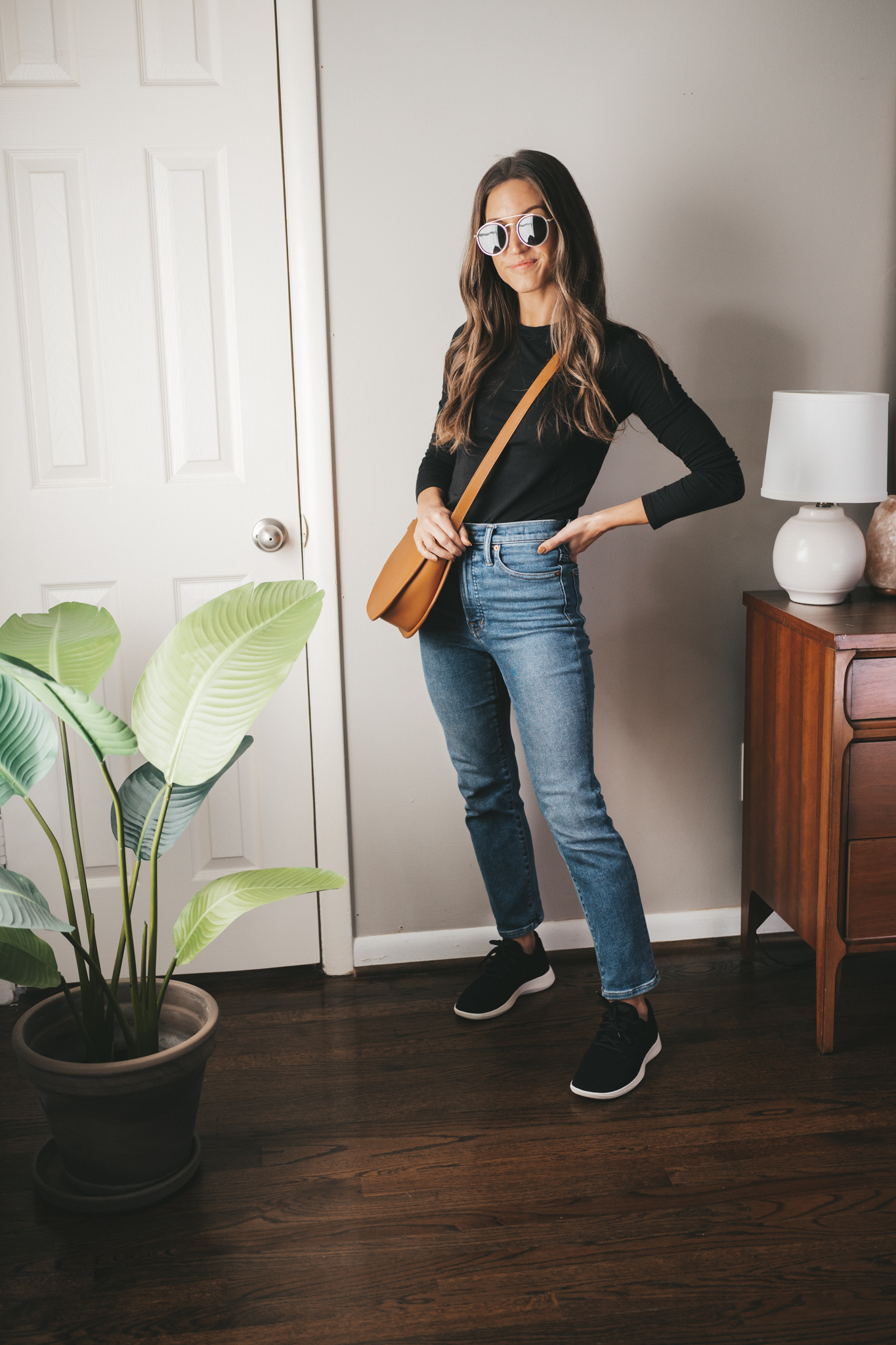 casual fall outfit idea with a black fitted long sleeved tee, mid-wash mom jeans, black sneakers, cognac crossbody bag, and round gold frame sunglasses