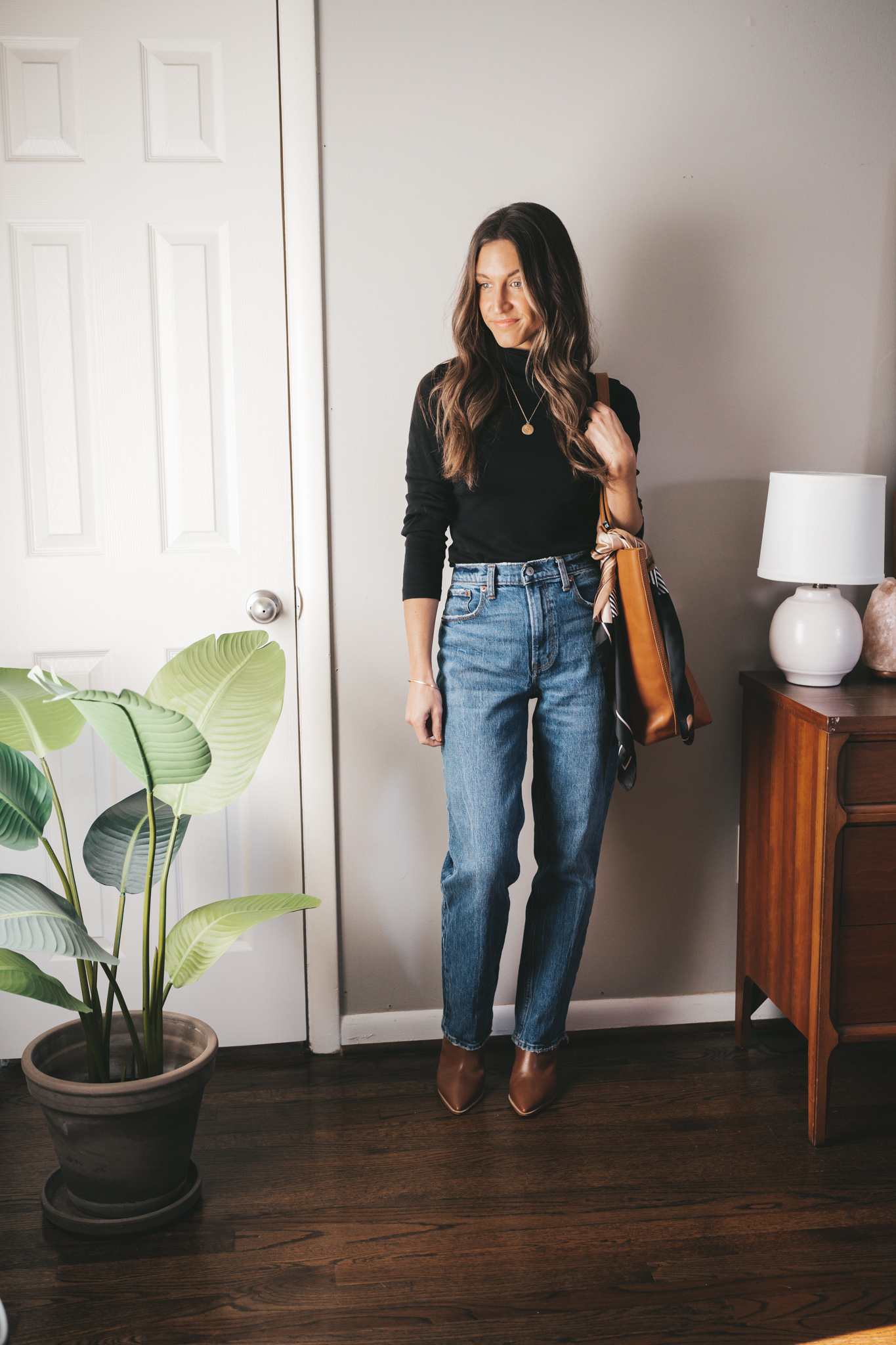 fall outfit idea with a black turtleneck, full length straight leg denim, cognac ankle boots, and a cognac tote bag with a silk scarf tied at the handle