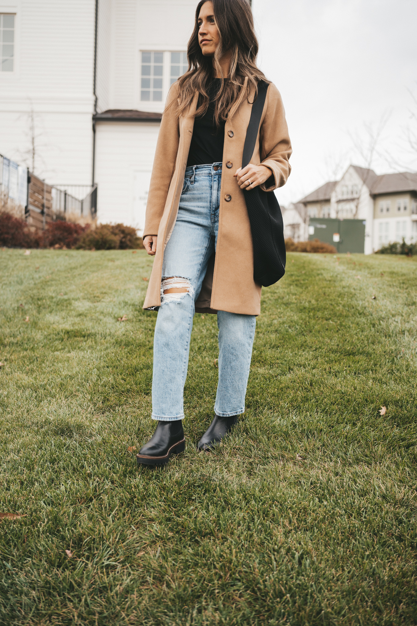 ashley standing on a small hill behind a white building wearing a black tee, camel coat, light wash distressed denim and black boots