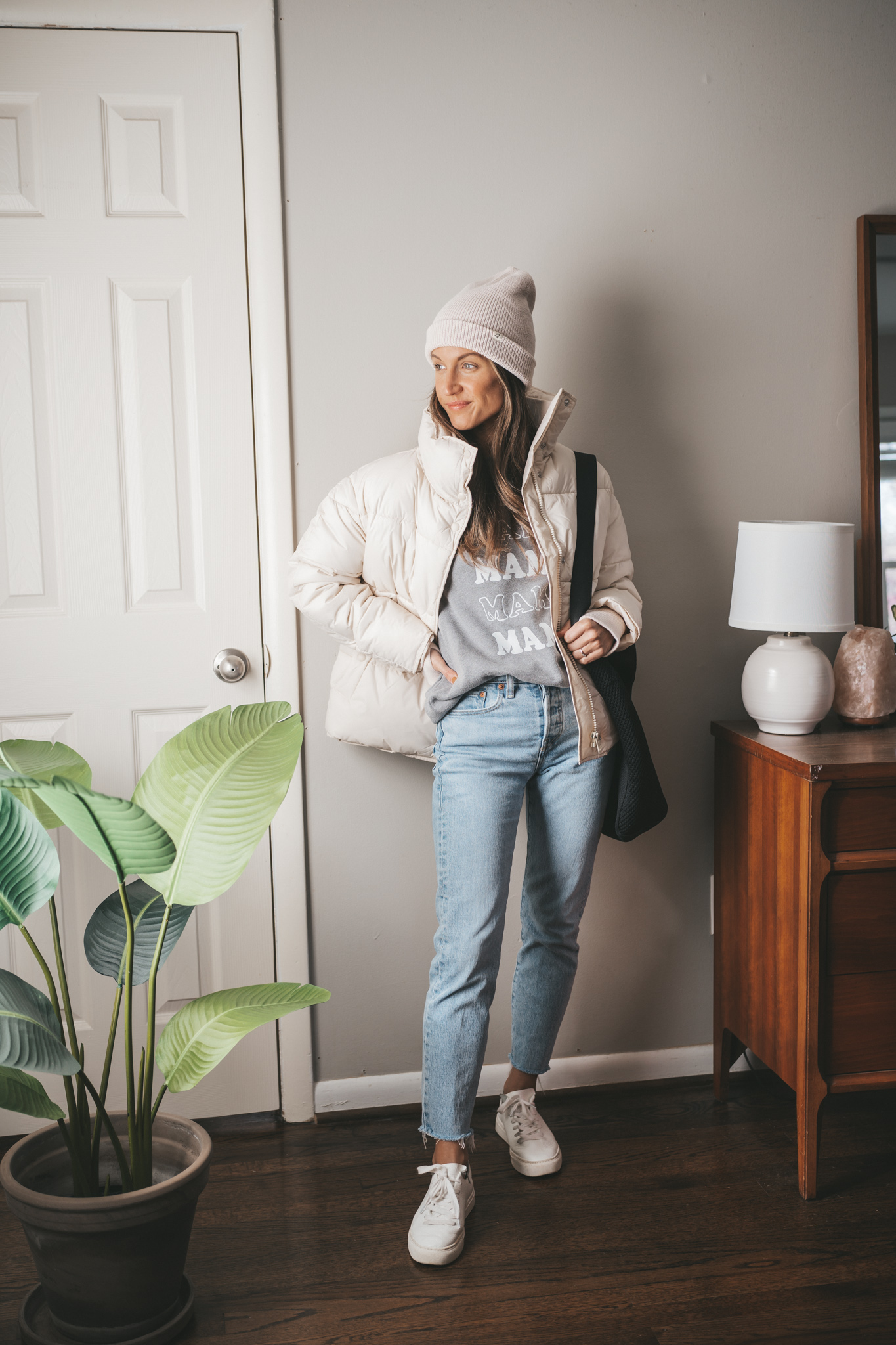 fall outfit idea with a graphic sweatshirt, light wash mom jeans, white sneakers, a white puffer coat, white beanie, and black crossbody bag