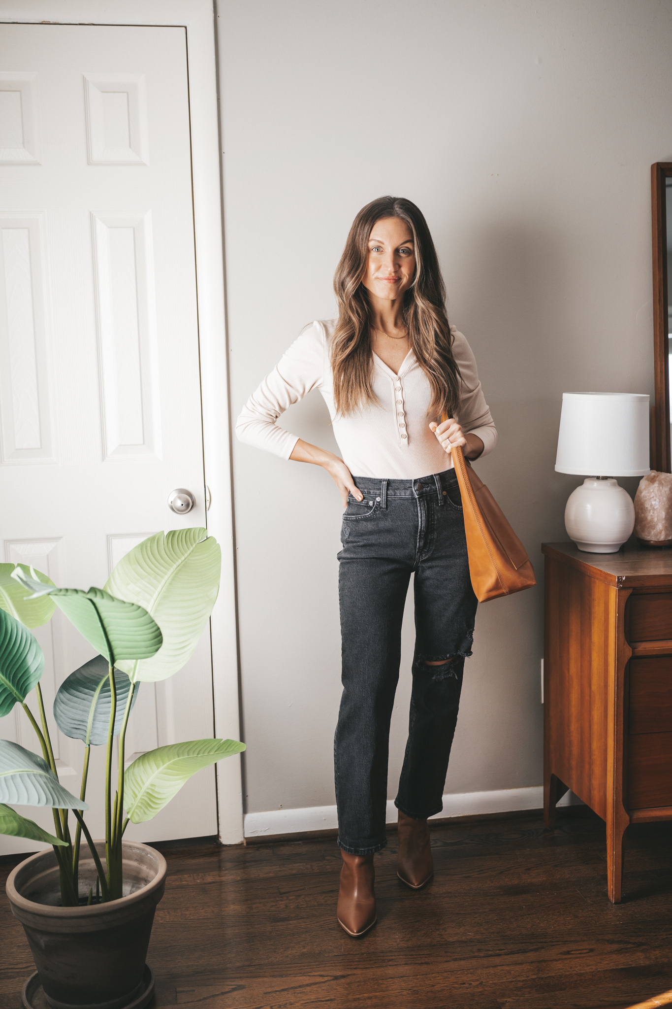 fall outfit idea with an ivory bodysuit, black straight leg denim, brown heeled ankle boots, and a cognac leather tote bag