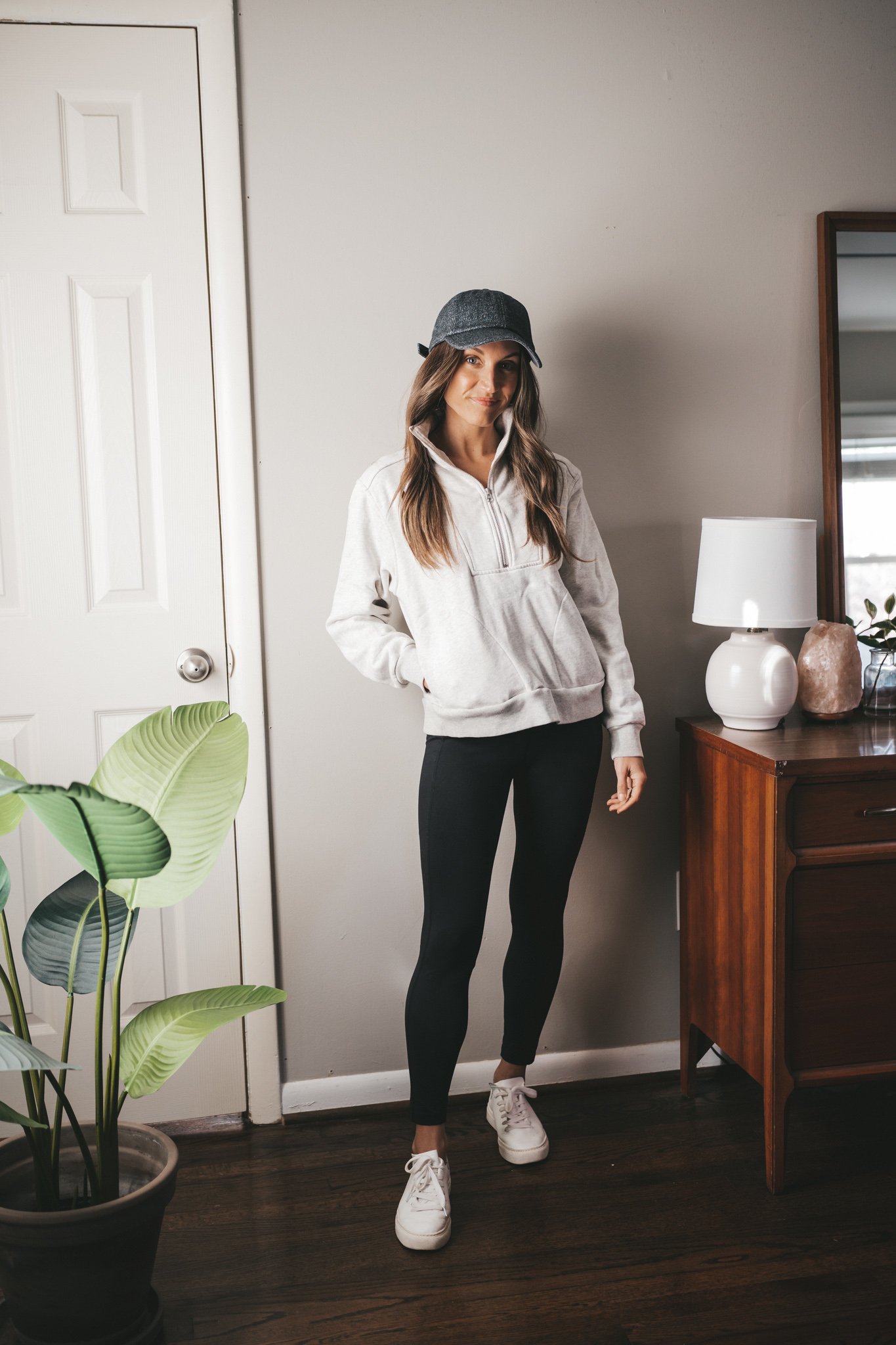leggings outfit idea with a light gray half zip sweatshirt, black leggings, white sneakers, and a dark gray baseball cap