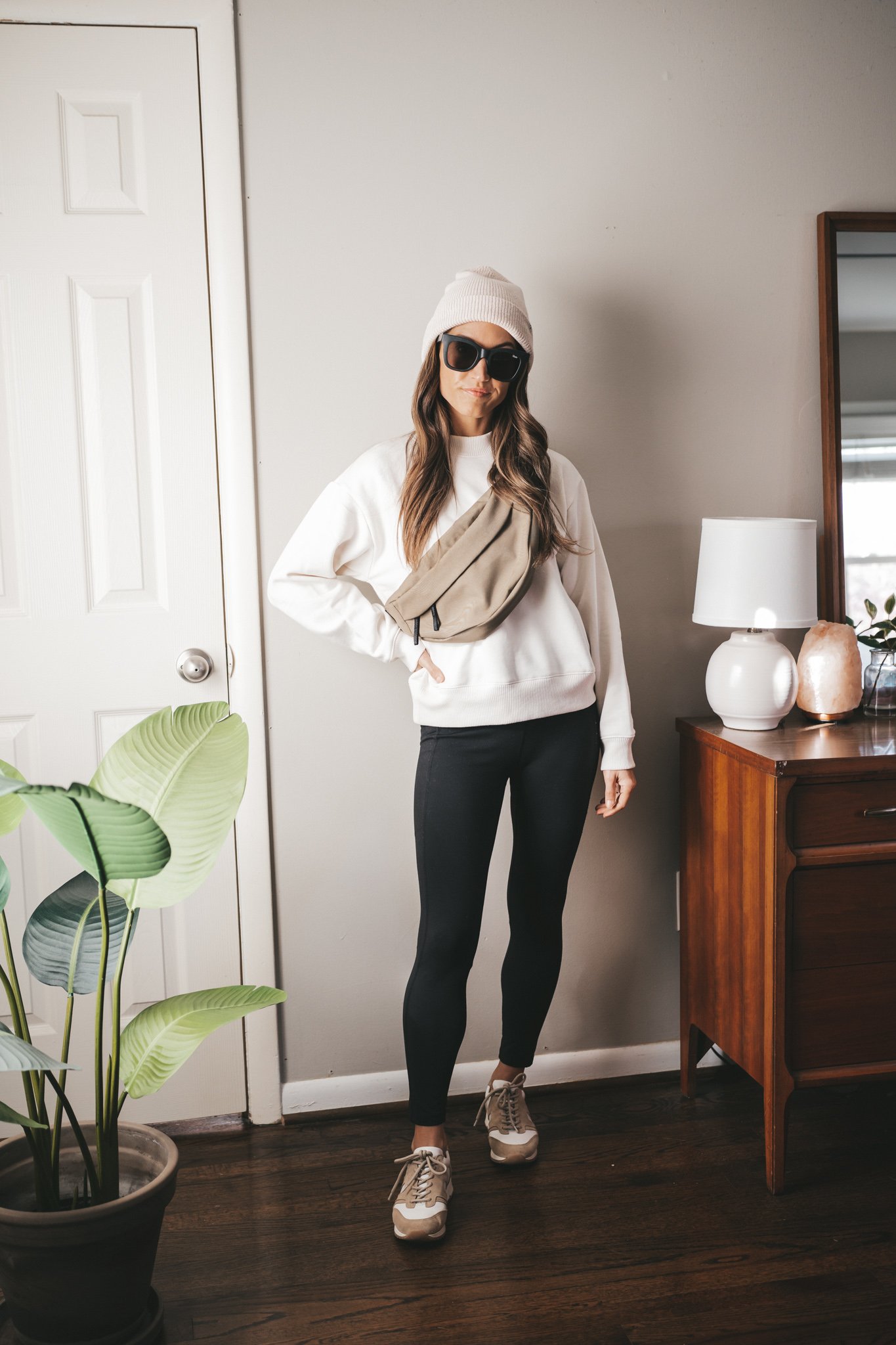 leggings outfit idea with a cream colored sweatshirt, black leggings, white and tan sneakers, an olive belt bag, black square frame sunglasses, and a cream colored beanie