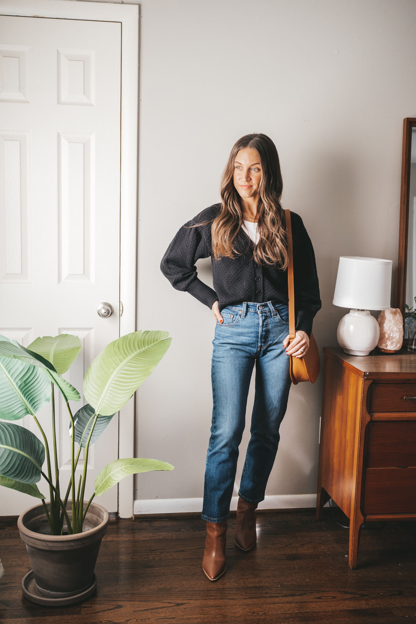 ashley wearing a white tee under a black button front cardigan with puff sleeves, mid-wash straight denim, brown heeled boots, and a cognac crossbody bag