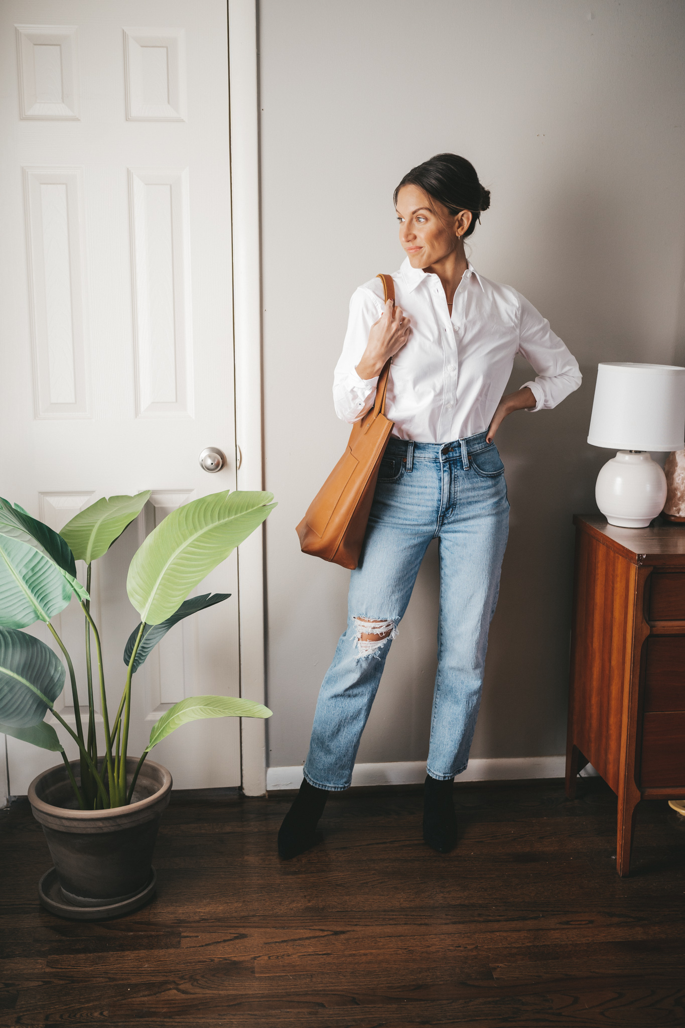 ashley wearing a white button down, light wash distressed straight jeans, black ankle boots, and a cognac tote bag
