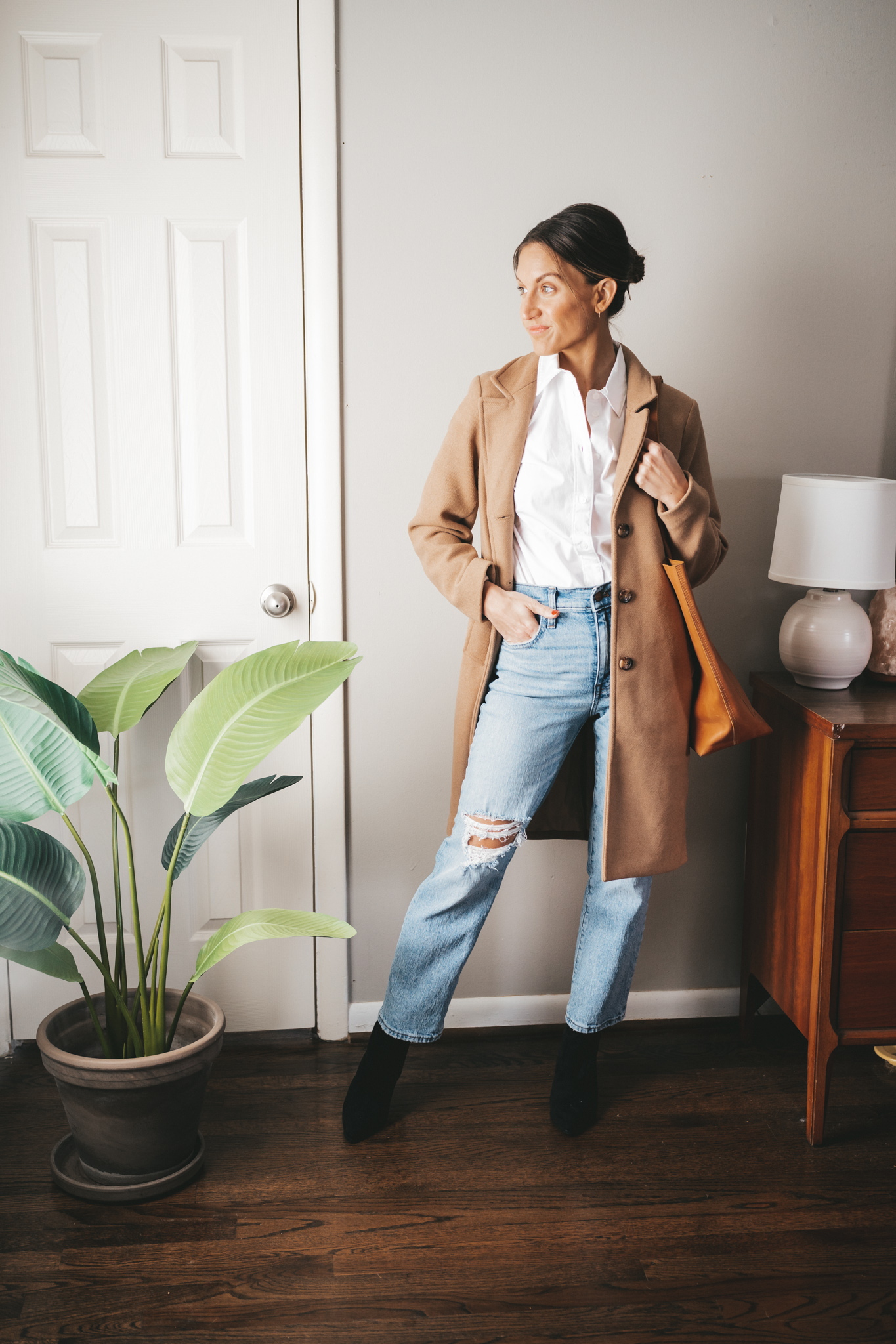 ashley wearing a white button down, camel coat, light wash distressed straight jeans, black ankle boots, and a cognac tote bag