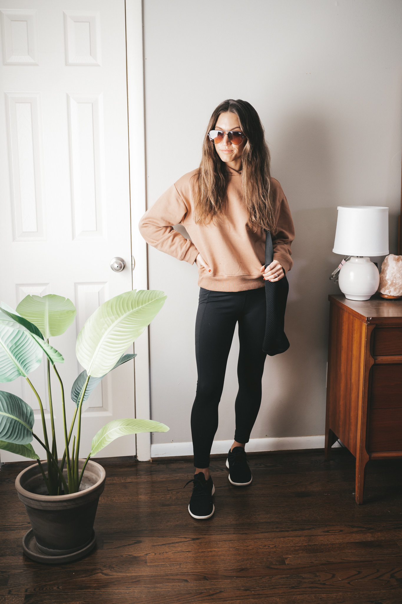 ashley wearing a brown hoodie, black leggings, black wool runners, and aviator sunglasses with a black mesh tote bag