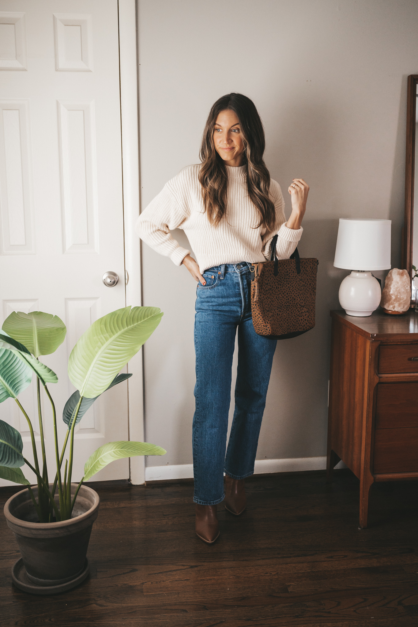 ashley wearing a chunky cotton cream colored sweater, mid-wash straight denim, brown heeled boots, and a leopard print tote bag