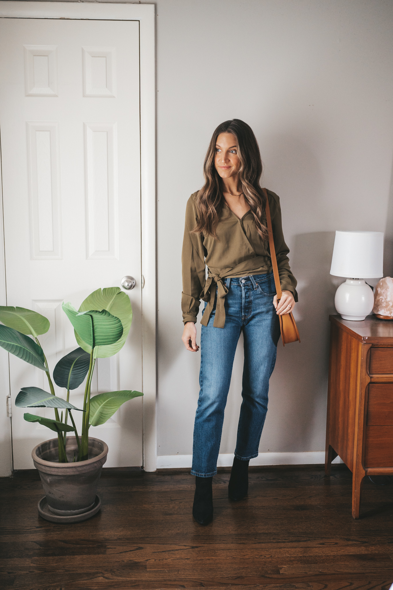winter outfit idea with an olive green wrap top, mid-wash straight denim, black suede heeled ankle boots and a cognac crossbody bag