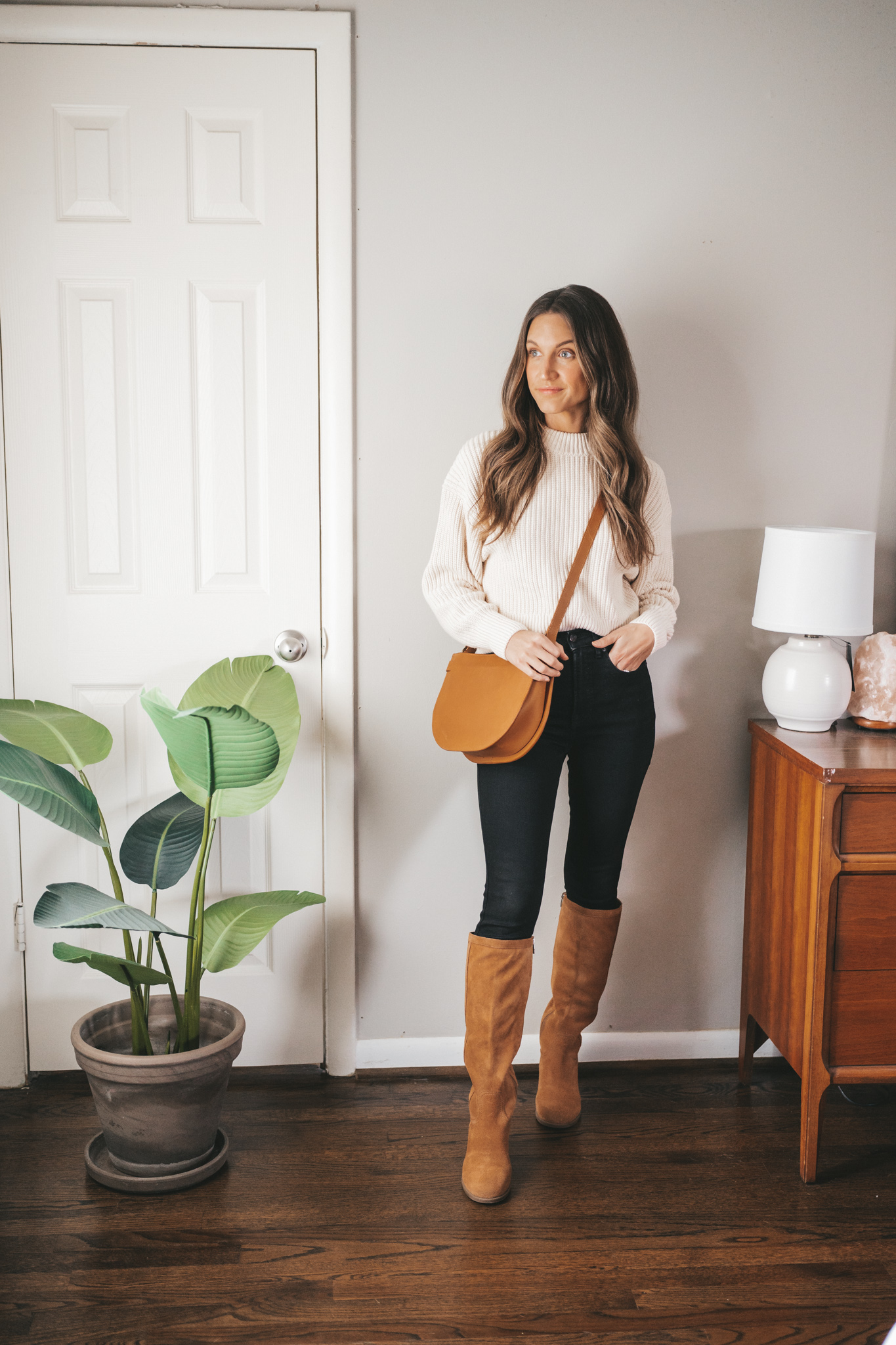 ashley wearing a Christmas brunch outfit with a cream colored chunky sweater, black skinny jeans, and cognac suede high knee boots, and a cognac crossbody bag