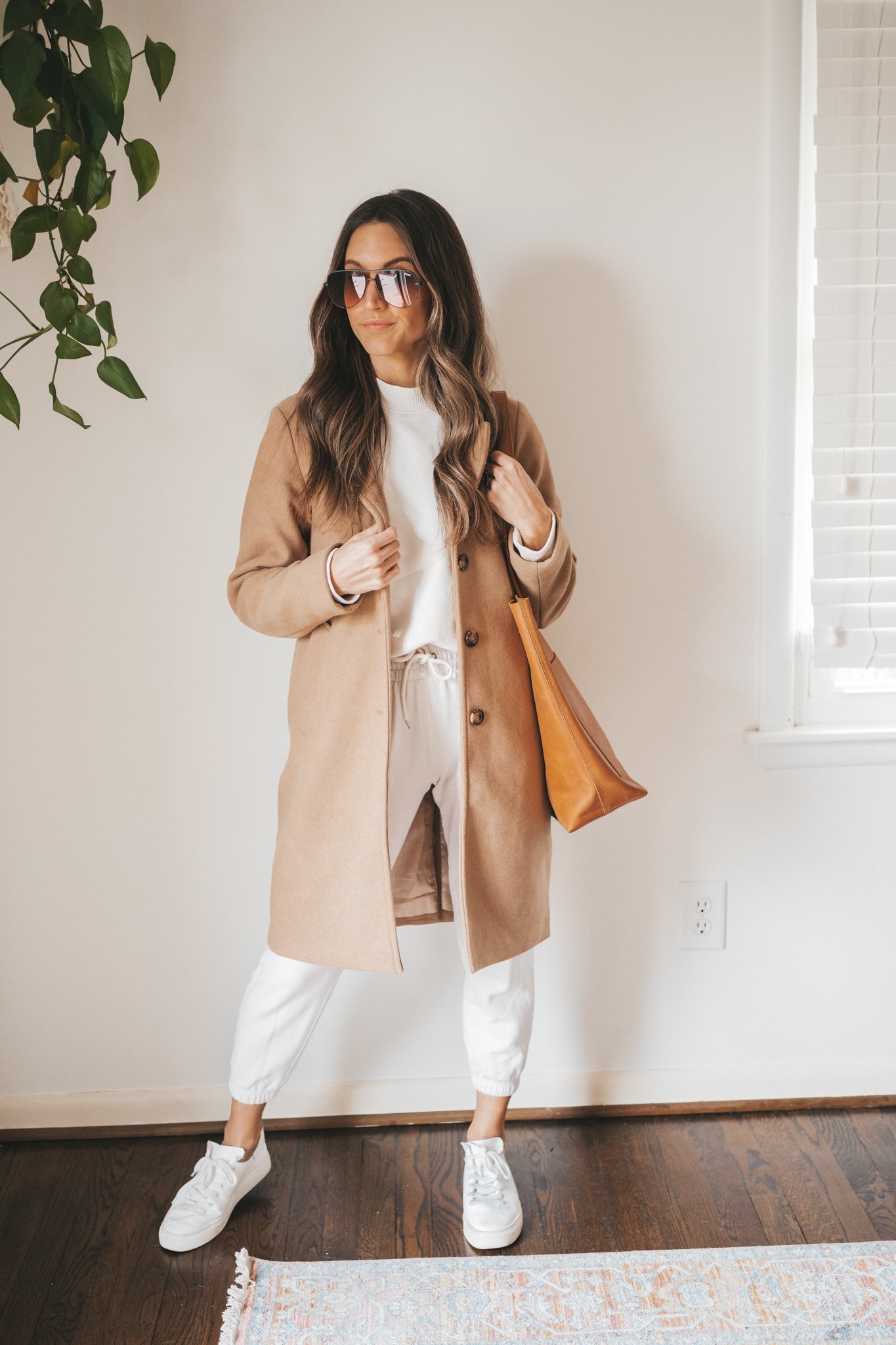 ashley wearing a cream colored sweatshirt, white joggers, a camel coat, white sneakers, a cognac leather tote bag and aviator sunglasses