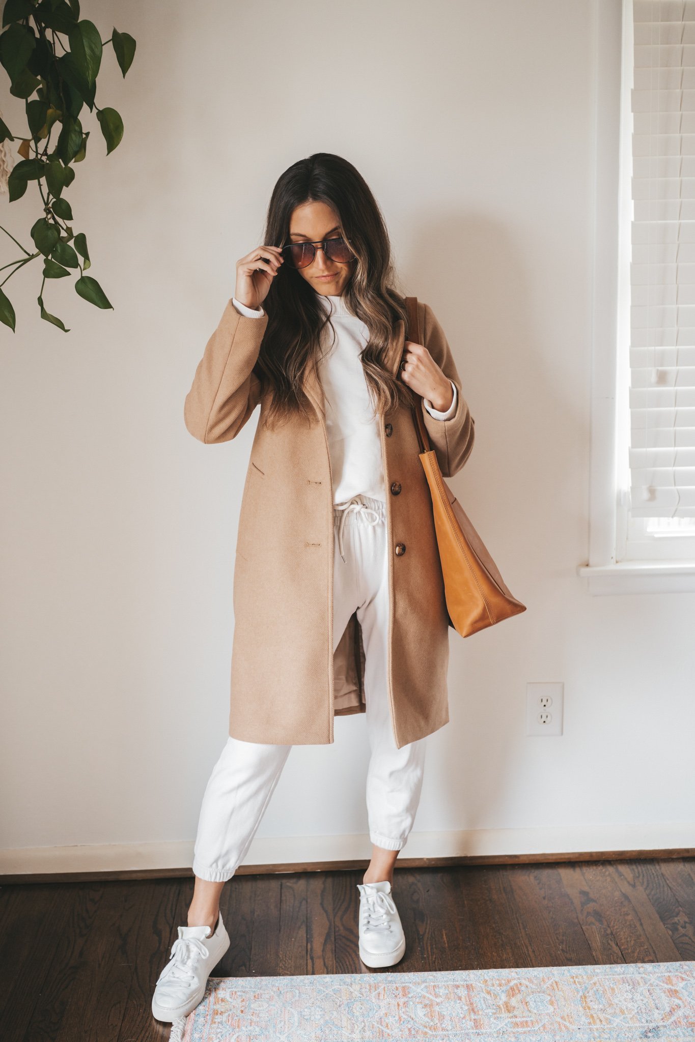 ashley wearing a cream colored sweatshirt, white joggers, white sneakers, a knee length camel coat, and sunglasses