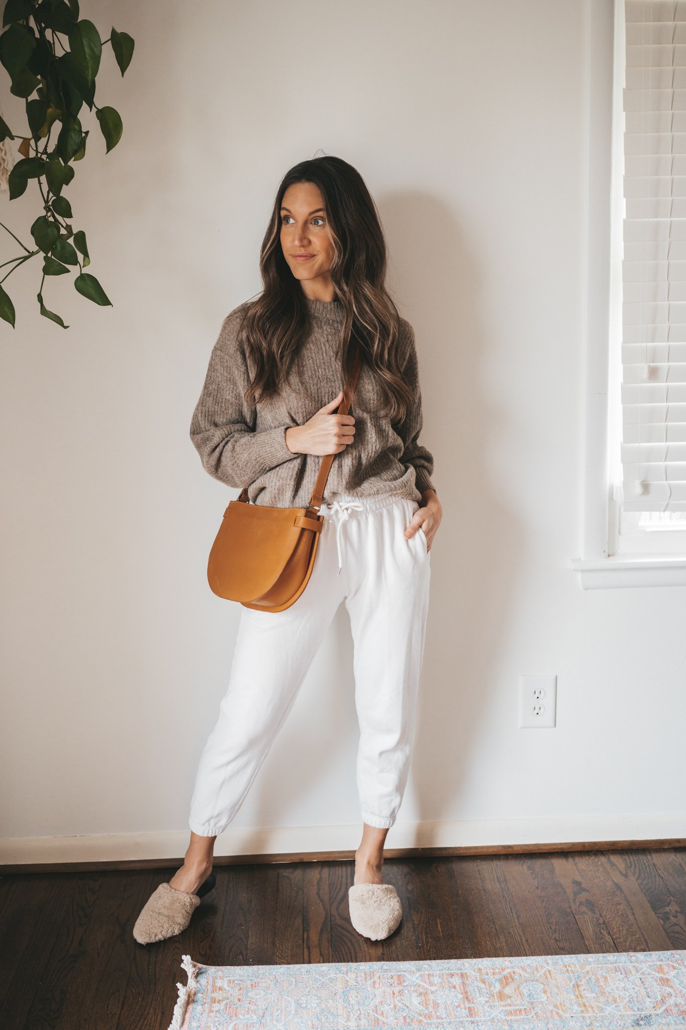 ashley wearing an oversized taupe sweater, white joggers, shearling mules, and a cognac crossbody bag