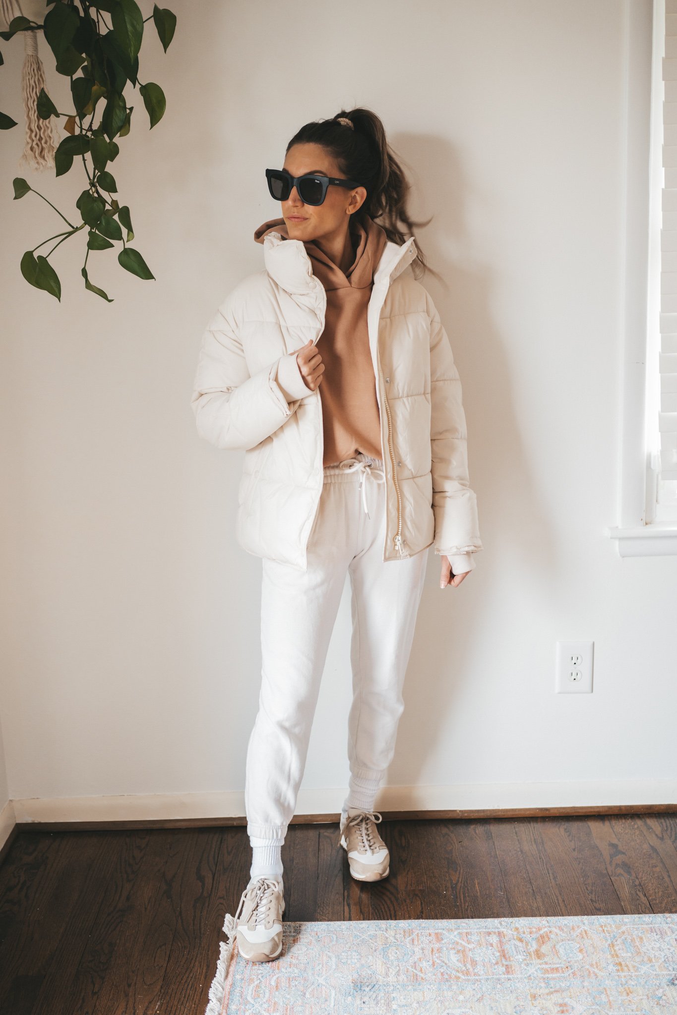 ashley wearing a brown hoodie sweatshirt, cream colored puffer, white joggers, white crew socks, and tan and white sneakers with black oversized square frame sunglasses