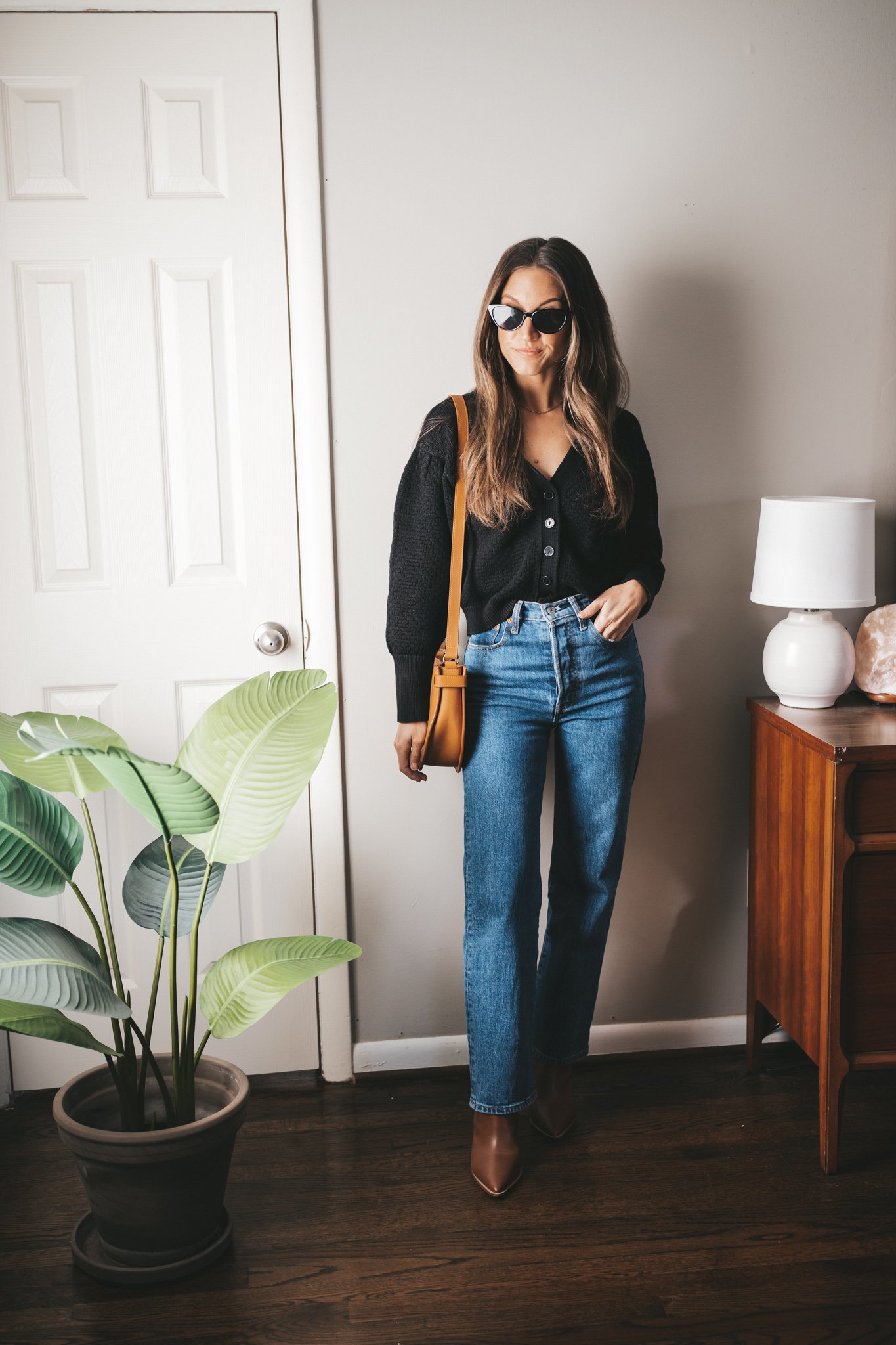 winter outfit idea with a black button front cardigan, straight leg medium wash denim, brown heeled ankle boots, and black cat-eye sunglasses
