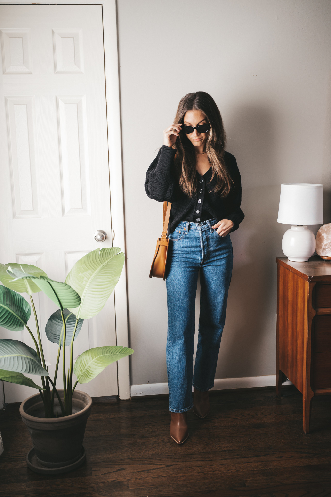 winter outfit idea with a black button front cardigan, straight leg medium wash denim, brown heeled ankle boots, and black cat-eye sunglasses
