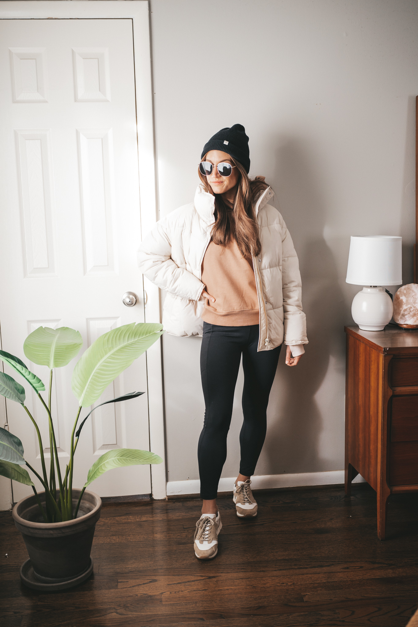 winter outfit idea with a brown hoodie, black leggings, white and tan leather sneakers, a white puffer coat, black beanie, and gold frame oversized sunglasses