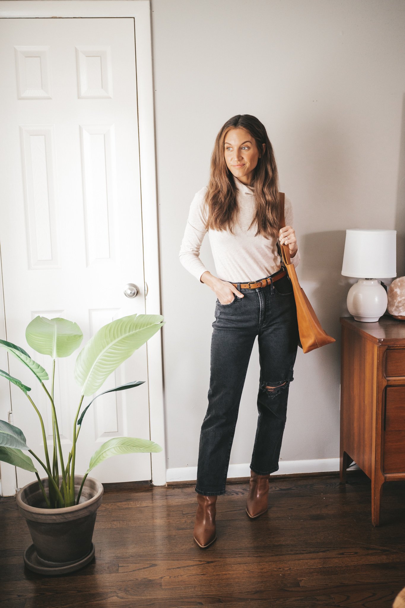 winter outfit idea with a beige turtleneck, washed black distressed denim, brown heeled ankle boots, cognac belt, and cognac tote bag.