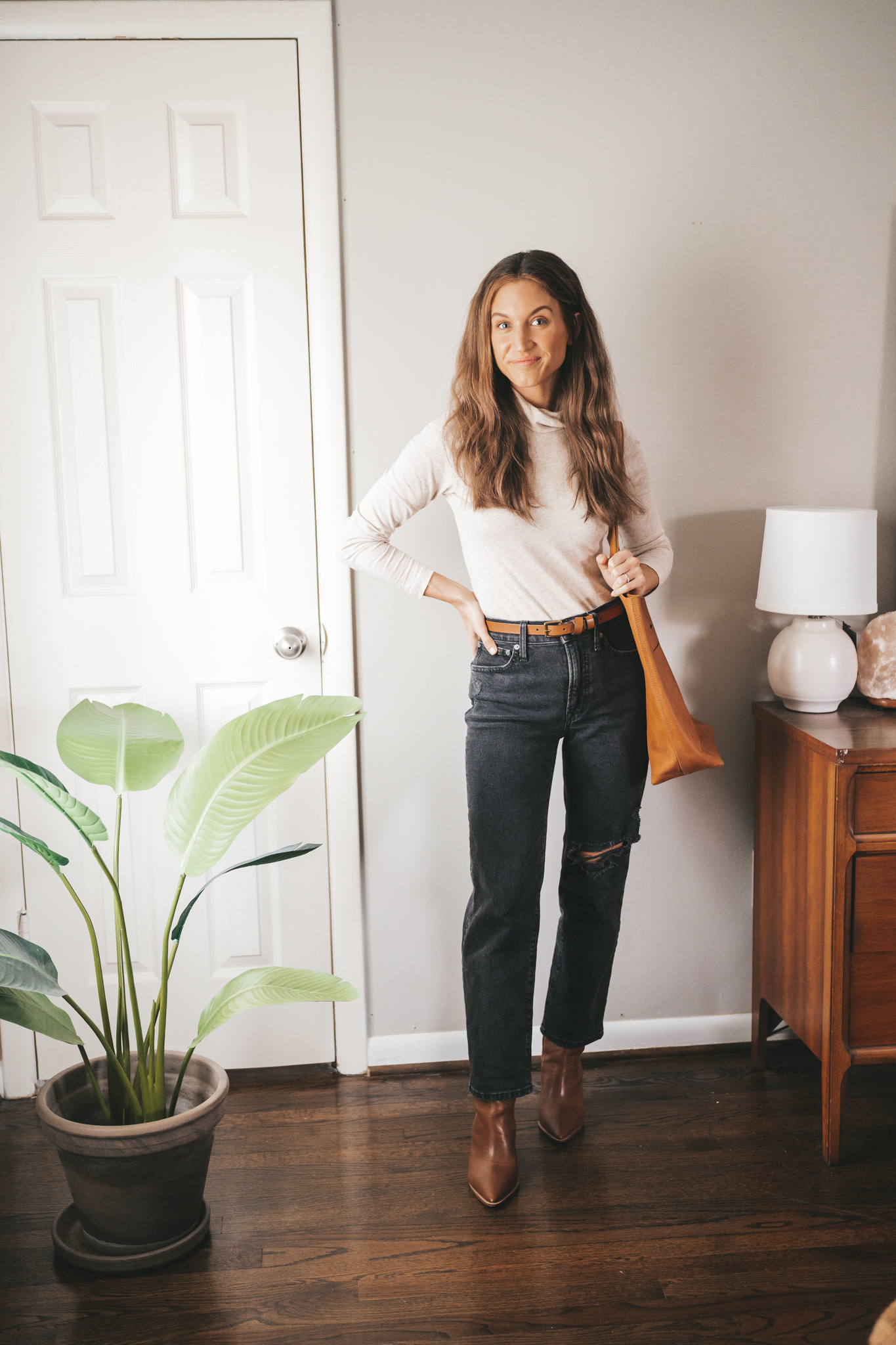 winter outfit idea with a beige turtleneck, washed black distressed denim, brown heeled ankle boots, cognac belt, and cognac tote bag.