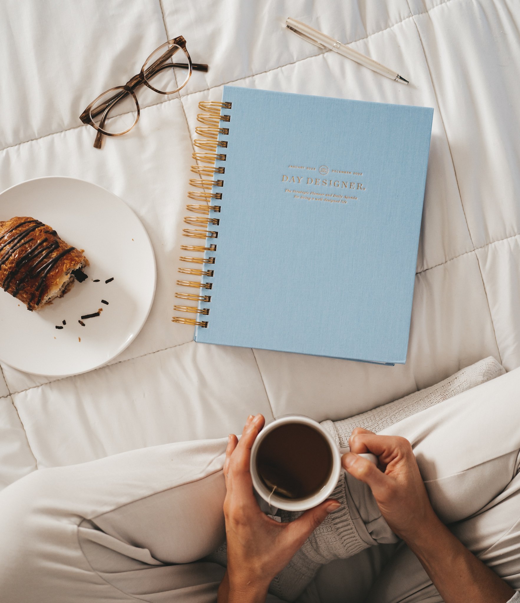 a white comforter with a cup of tea, a chambray blue Day Designer Planner, glasses, a chocolate croissant, and a silver pen